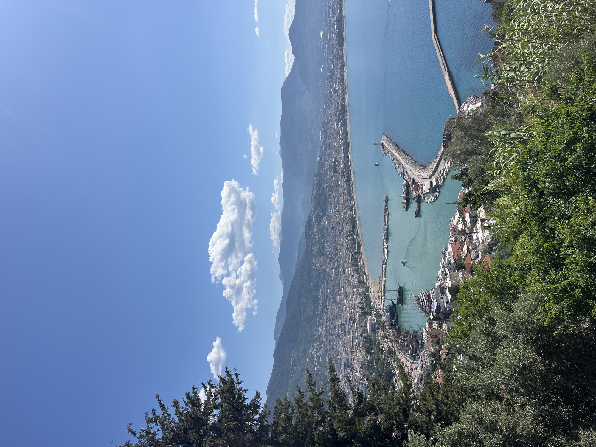 The image is a high-angle, daytime shot of Alanya, Turkey, taken from an elevated position, potentially from the Alanya Castle. The shot is composed of the following elements:
**Environment:** The environment consists of a clear blue sky with scattered cumulus clouds, a mountain range extending along the coast, a densely built city that runs along the coast, and the Mediterranean Sea. The foliage suggests a warm, sunny day.
**Objects:** Dominating the scene is the coastline, with Alanya's urban area stretching along it. A long pier or jetty extends into the sea, where numerous boats are docked. The city is a mix of buildings, which appear to be a combination of residential and commercial structures. The mountains form a backdrop to the city. Trees and shrubbery are visible in the foreground.
**Location:** Alanya, Turkey, is a coastal city with a harbor. The presence of the mountains, the cityscape layout, and the harbor detail indicate the shot is likely taken from the high ground, probably near Alanya Castle.
There are no people visible in the image. No actions or interactions are discernible other than the natural landscape elements and urban elements combining.
