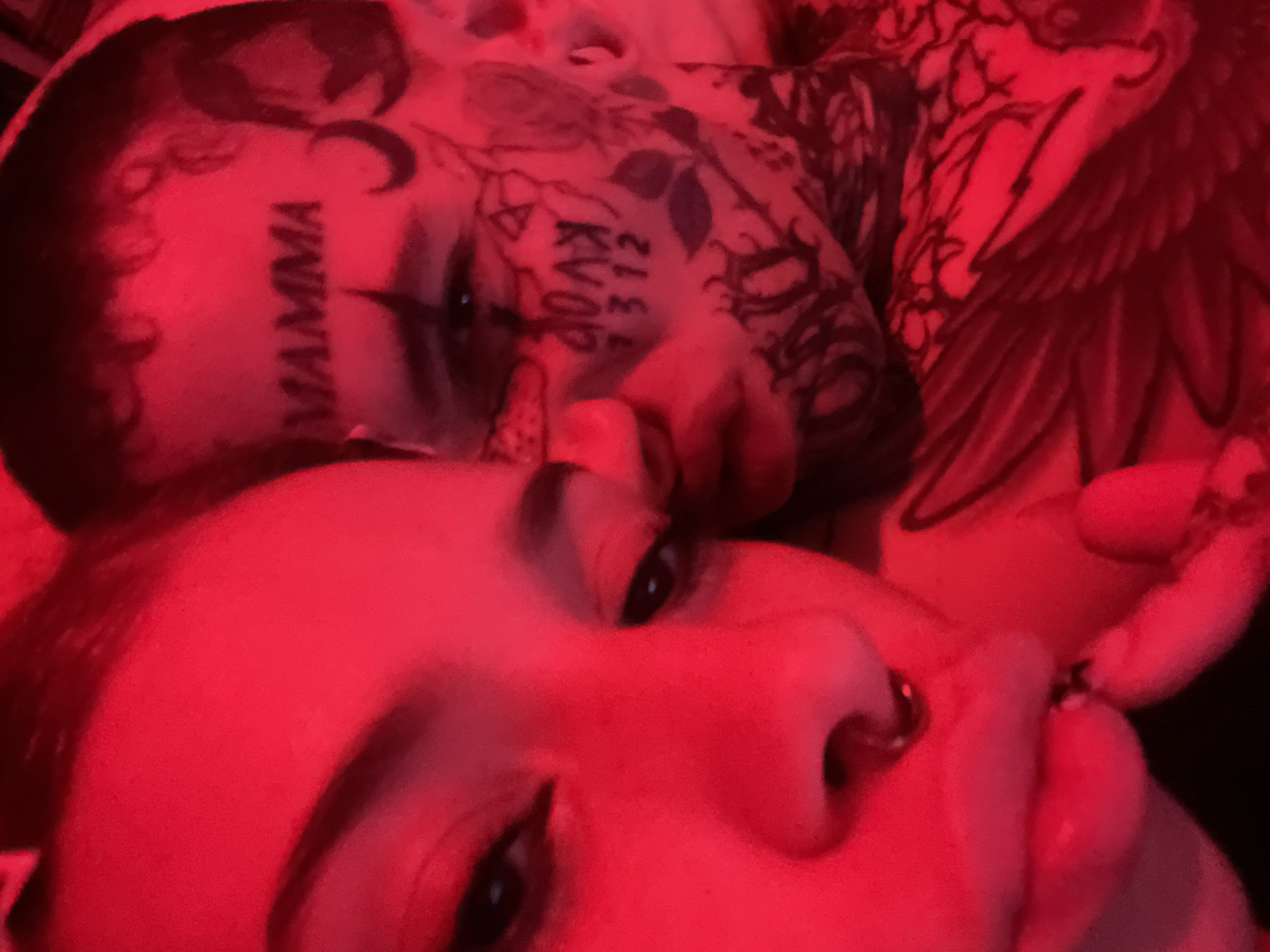 Two individuals are visible in a close-up shot, illuminated by a strong red light. The person in the foreground, likely female, has fair skin and dark eyes, and a nose piercing. The second person, positioned behind and slightly above, appears male with extensive tattoos on their face and upper body. Tattoos are visible on their forehead which reads "MAMMA" with a crescent moon above it, and on their cheek which reads "KVOD 312". The background is indistinct due to the low light and close proximity to the subjects. There is no information in the photograph to determine that it was taken in Buenos Aires, Argentina.