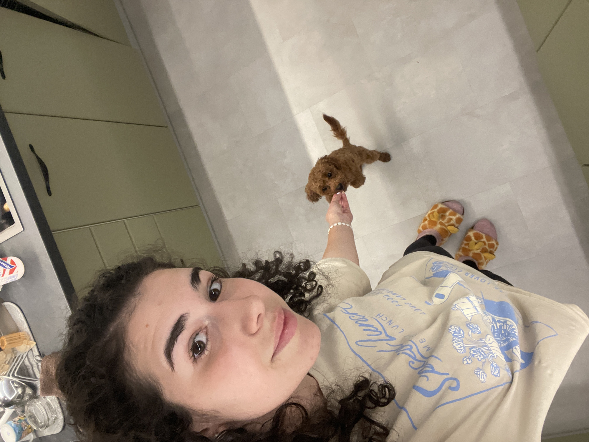 A woman with curly hair is taking a selfie in a kitchen. She is wearing a light-colored t-shirt with blue writing and a graphic design, and giraffe-patterned slippers. Her left arm is extended, offering something to a small, brown dog. The kitchen has light-colored tiled flooring and cream-colored cabinets with dark handles. The location is presumed to be Sofia, Bulgaria, though there are no explicit landmarks to verify.