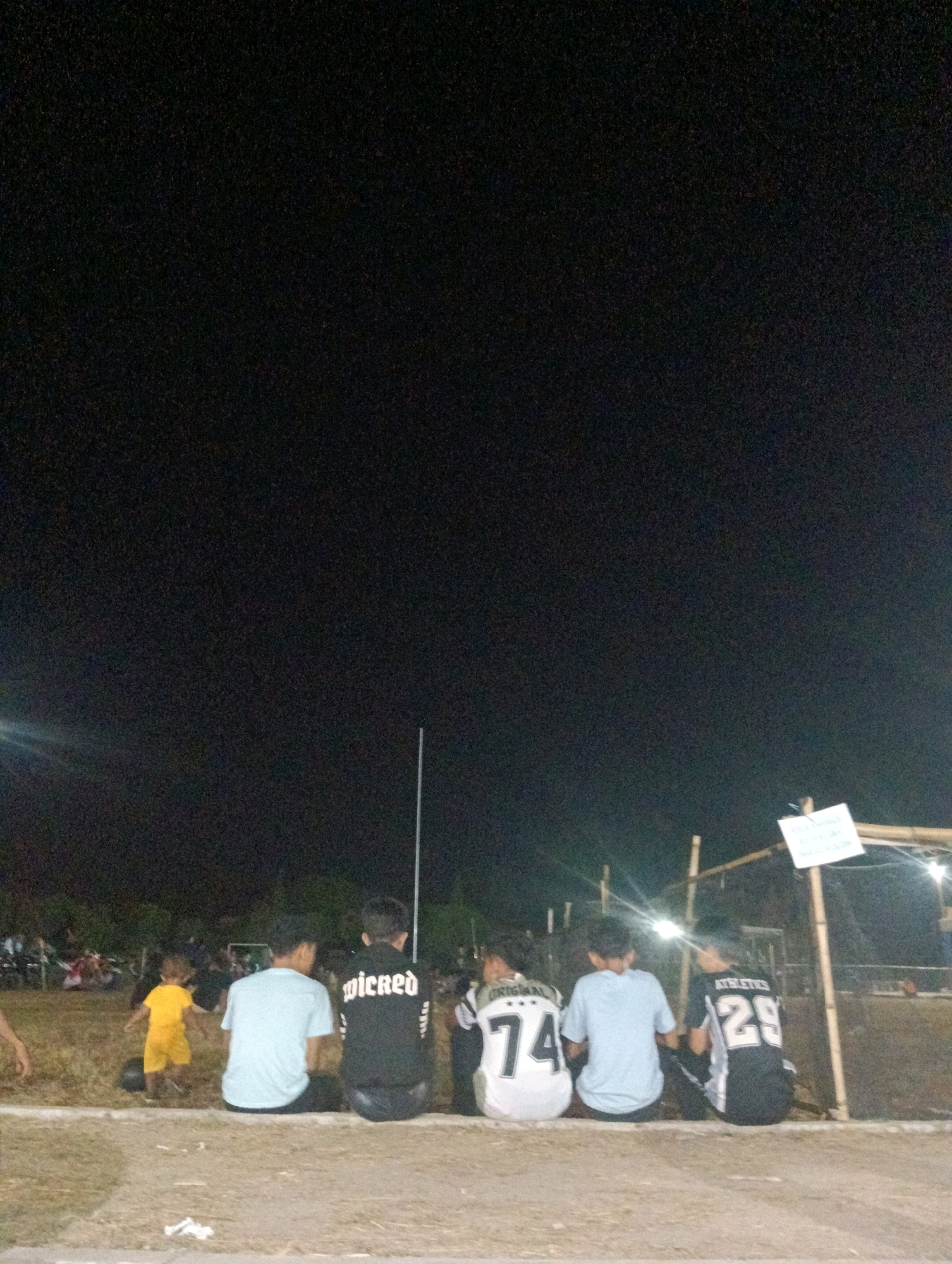 The image depicts a nighttime scene in Pematang Jaya, Indonesia, likely at a sports field or gathering area. Four young males are seated on a low barrier, facing away from the camera. The individual on the far left is wearing a light blue t-shirt and dark pants. Next to him, a person in a black t-shirt with white lettering that reads "wicked" is visible. To their right, another person wears a white jersey with the number "74" and the word "ORIGINAL" printed on it, paired with light blue shorts. The individual on the far right is dressed in a black jersey with white lettering and the number "29" on the back, along with the word "ATHLETICS." In the background, a small child in a yellow outfit is running on a grassy area. Various indistinct figures and structures, possibly spectators and a makeshift shelter, are present further in the background, illuminated by artificial lights. The overall environment appears to be an outdoor event or recreational space at night, with elements suggesting a community gathering.