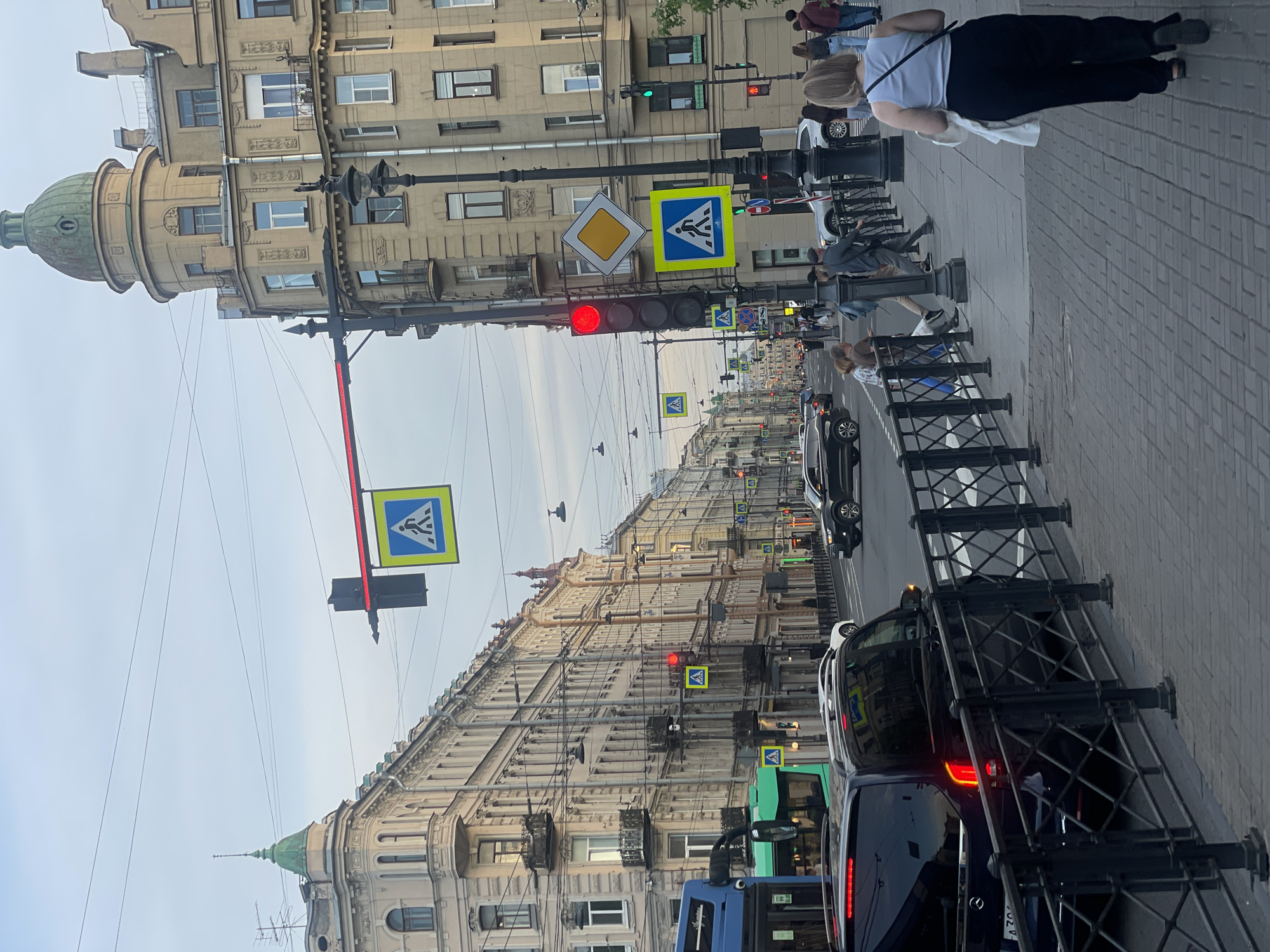 This detailed image captures a street scene in the city of Saint Petersburg, Russia, likely on Ligovsky Prospekt, characterized by its distinctive architecture. The time of day appears to be late afternoon or early evening under an overcast, light gray sky. In the foreground, a multi-lane asphalt road extends into the distance, flanked by sidewalks. On the immediate left, the rear of a dark-colored Mercedes-Benz minivan or SUV is visible, its red taillights illuminated. A partially visible Russian license plate, featuring characters like "A 70," is affixed to its rear. A black metal fence with a diamond pattern separates the vehicle from the sidewalk. The sidewalk on the right is made of interlocking pavers. A woman with light-colored hair, wearing a light blue sleeveless top and dark pants, walks away from the viewer, carrying a white bag in her left hand. Further along the sidewalk, a man in a dark cap and gray jacket walks past a black, ornate lamppost. Other pedestrians, including a man in a brown shirt and a woman in a floral top, are visible closer to the street. The street is lined with numerous multi-story buildings showcasing ornate European architectural styles. On the left, a prominent light-colored corner building features a green conical spire. Further down, a continuous row of similar buildings with balconies and decorative facades extends along the street. On the right, a large, ornate corner building dominates the scene, characterized by a weathered, greenish-copper dome (cupola) at its apex and intricate detailing on its facade. Overhead, a network of electrical wires, presumably for trolleybuses or trams, crisscrosses the sky. Multiple traffic lights are active: a prominent red light is visible on a pole on the right, and another red horizontal light bar is suspended over the street. Other green and red traffic lights are visible further down the road. Several blue square signs with a white triangle and black pedestrian symbol (indicating a pedestrian crossing) are suspended over the road and mounted on poles. A yellow diamond-shaped sign with a white border (indicating a priority road) is also mounted on a pole on the right. Vehicles are present on the road. In the mid-ground on the left, a blue articulated bus and a green electric bus (likely a trolleybus) are visible. Further down the street, various cars, including a white SUV, a silver sedan, and a black SUV, are in traffic, with the
