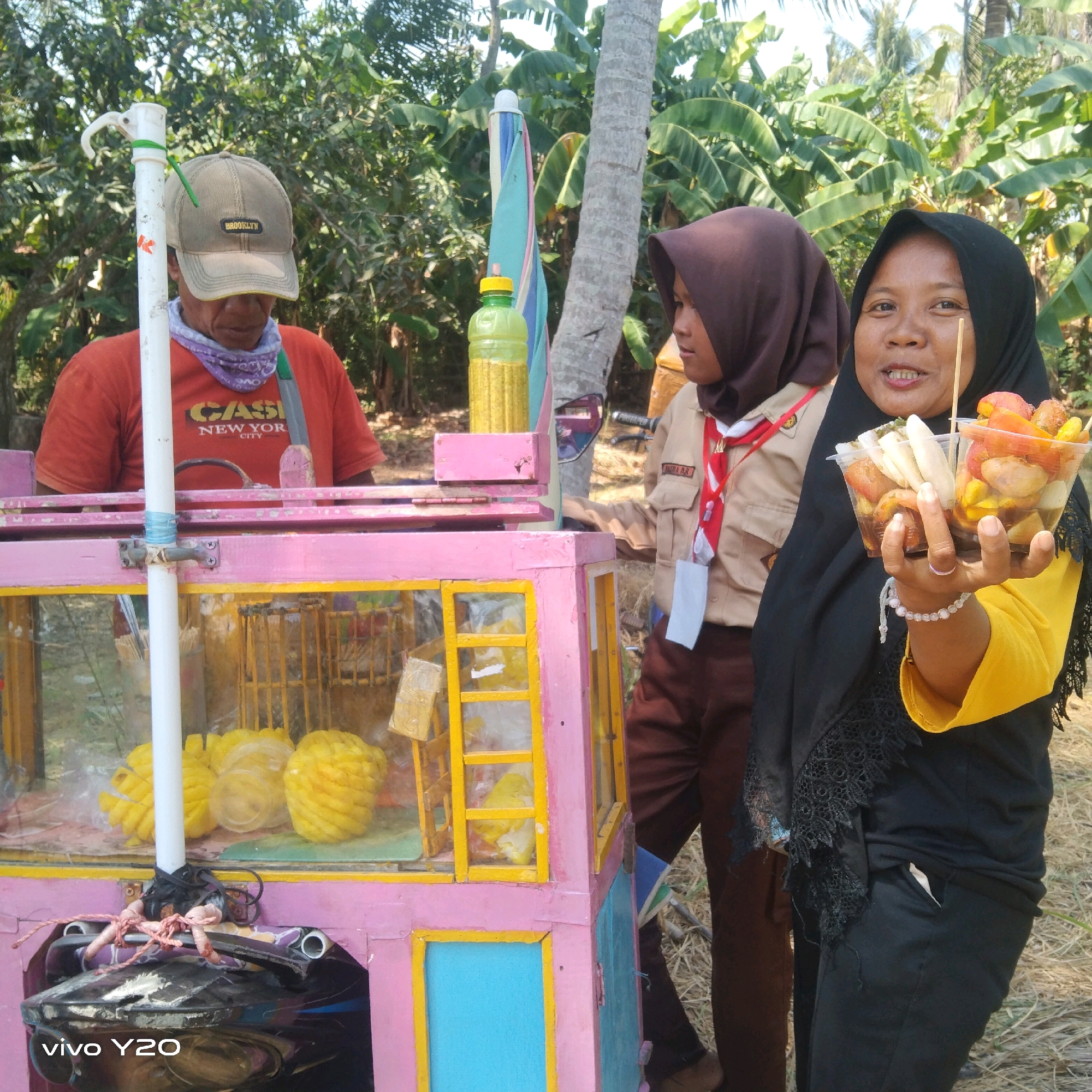 In Karawang, Indonesia, a street vendor selling food stands beside a bright pink food cart. The cart is adorned with yellow trim and a blue drawer. Inside the cart, sliced pineapples are displayed. To the right of the cart, a woman in a black hijab and yellow top holds up two clear plastic cups filled with various fried snacks. Next to her, a young girl wearing a brown scout uniform with a red and white sash stands. Behind them, lush green banana leaves and palm trees form a dense backdrop. The ground is covered with dry grass and twigs. A white pole, possibly a flag pole or part of the cart's structure, is positioned prominently in the foreground, secured to the cart with pink and black ropes. The scene is illuminated by natural daylight, suggesting it is daytime.