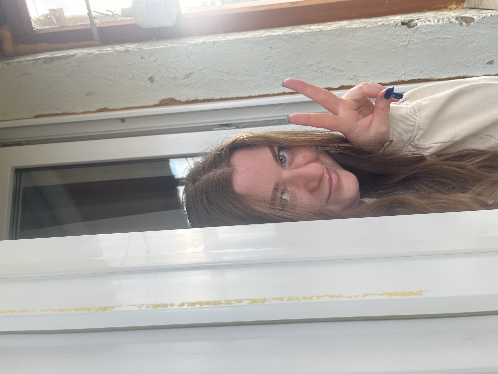 A woman with light brown hair and blue nail polish gives a peace sign in front of a window with a white frame and a mottled white wall above. Yellowish residue is visible on the window frame. The location is Раздолье, Russia.