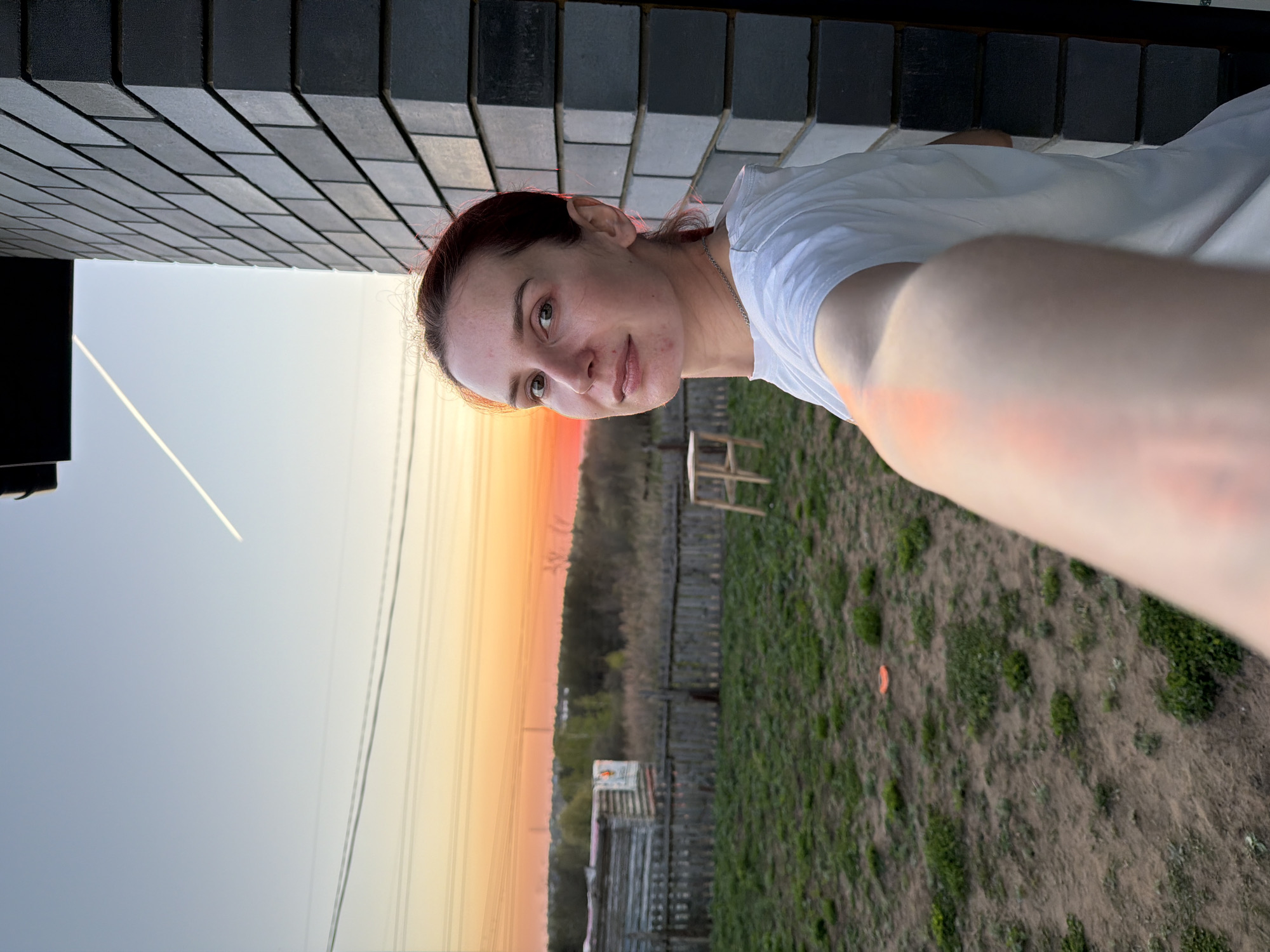A person is holding a camera up to take a selfie. The location appears to be Chaikovsky, Russia, based on the Cyrillic alphabet and the general architecture. The selfie is taken near a brick wall. The background features a sunset or sunrise with power lines, a fence, a patch of green vegetation, and a small wooden stool. The person in the selfie appears to be wearing a white t-shirt.