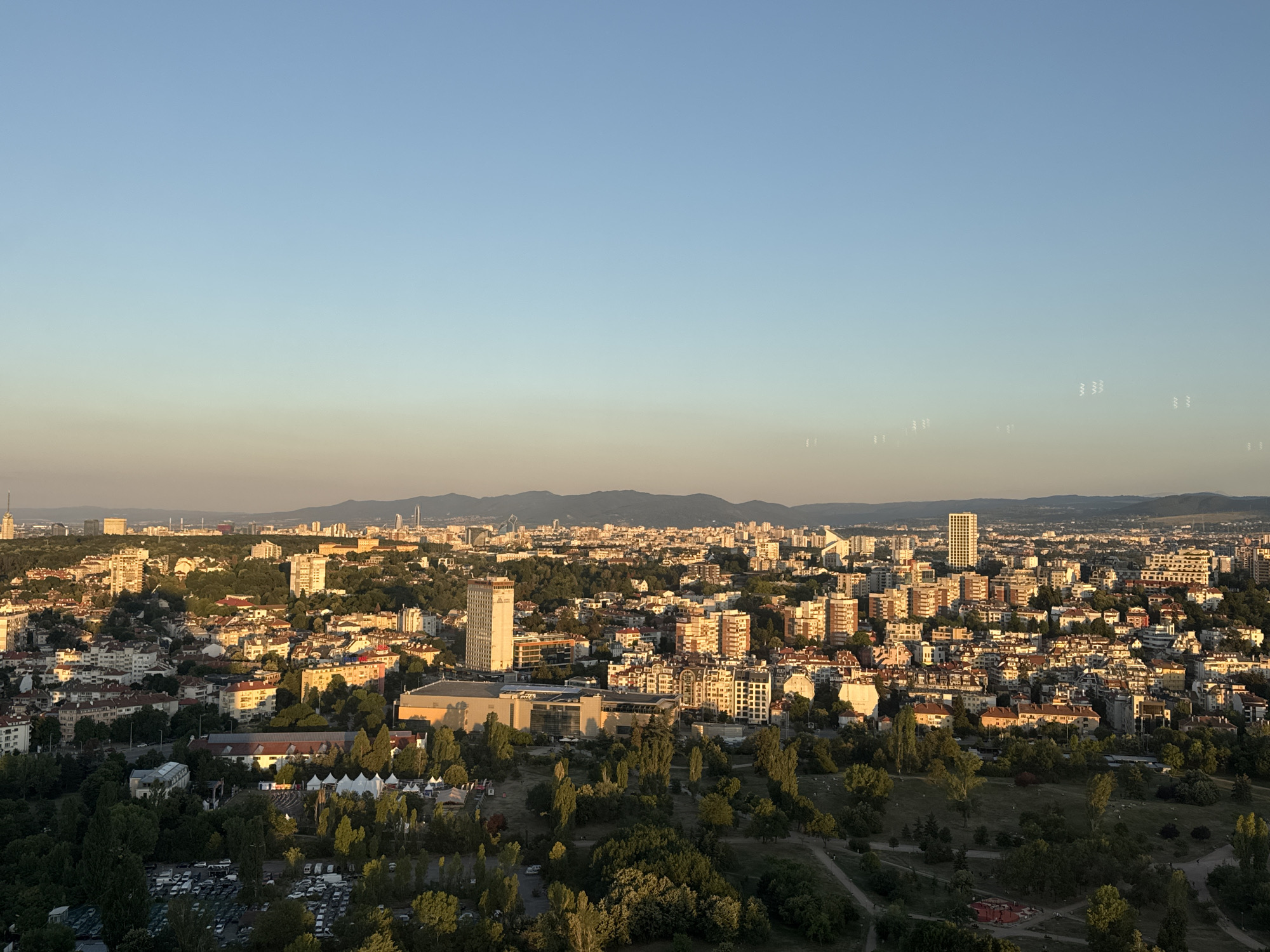 This is an aerial panoramic view of Sofia, Bulgaria, likely taken from a high vantage point. The image captures a sprawling urban landscape with a mix of residential buildings, commercial structures, and green spaces. The golden hour sunlight casts long shadows and bathes the city in a warm glow, suggesting it is either sunrise or sunset.
In the foreground, the scene transitions from a densely packed area of apartment buildings to a more park-like setting with mature trees and open grassy areas. A large parking lot filled with numerous cars is visible on the lower left. There are also several white tent-like structures, possibly indicating an event or market, and playground equipment.
The mid-ground is dominated by a dense concentration of apartment buildings of varying heights and architectural styles, painted in a variety of colors, predominantly light hues. A prominent tall, slender building with a distinctive modern design stands out among the other structures, with a logo visible on its upper section. Adjacent to it is a large, rectangular building with a glass facade and a light-colored roof, possibly a cultural or commercial center.
The background reveals a wider view of the city extending towards a distant mountain range, silhouetted against the pale blue sky. The sky itself exhibits a gradient of color, transitioning from a clear blue at the top to a warm, hazy yellow near the horizon, consistent with the time of day. Scattered white, wavy shapes are visible in the sky, which could be atmospheric effects or light refractions. The overall impression is of a vibrant city at the close of day, with its urban fabric interwoven with nature.