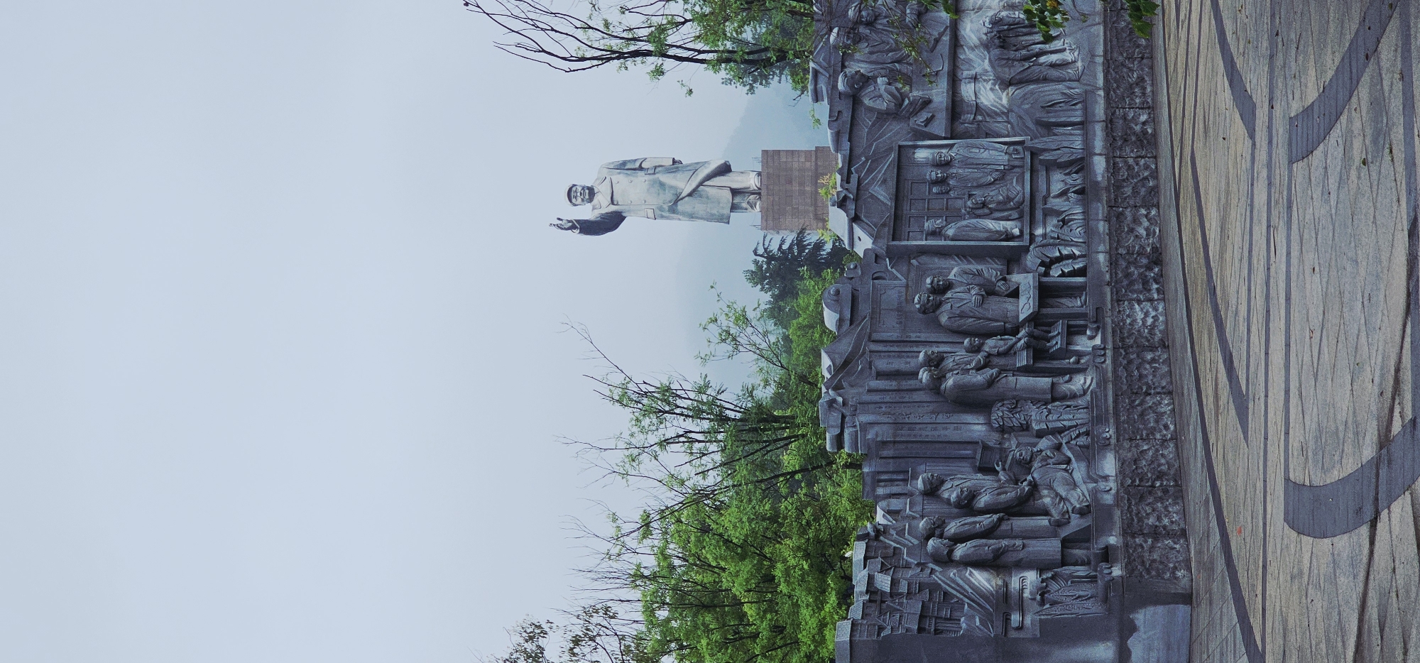 The image depicts a statue of a man in a long coat and cap, with his right arm raised. He stands on a raised platform in front of a gray, sculpted relief that features numerous figures in various poses. The relief is set against a backdrop of lush green trees and a misty, mountainous landscape. The foreground shows a paved area with geometric patterns. This monument is located in the Chongqing High-tech Zone, China.