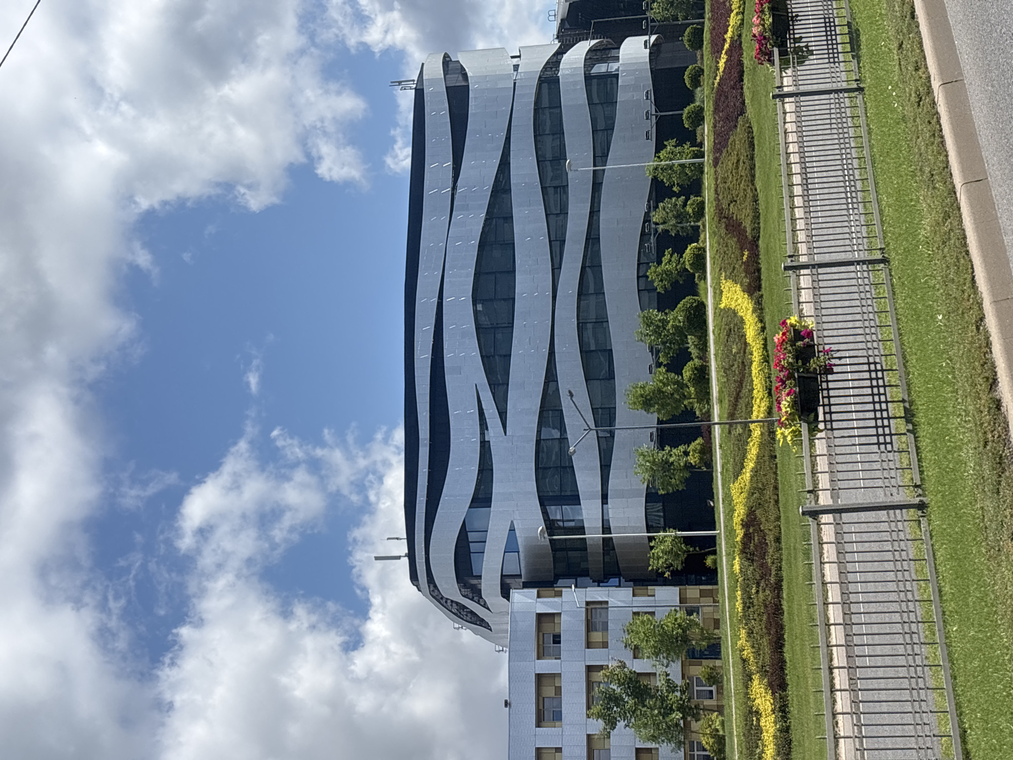 The image depicts a modern building with a distinctive wavy facade, located in Minsk, Belarus. The building is several stories high and features a metallic exterior with horizontal, undulating bands. Adjacent to this structure is a less ornate, lighter-colored building with visible windows. In the foreground, a neatly maintained green lawn is separated from a road by a gray metal fence. Beyond the fence, a strip of land features patterned landscaping with yellow and red flowers and dark green shrubs. The sky above is blue with scattered white clouds. There are no people visible in the image. The scene suggests an urban or suburban environment, likely a business or institutional complex.