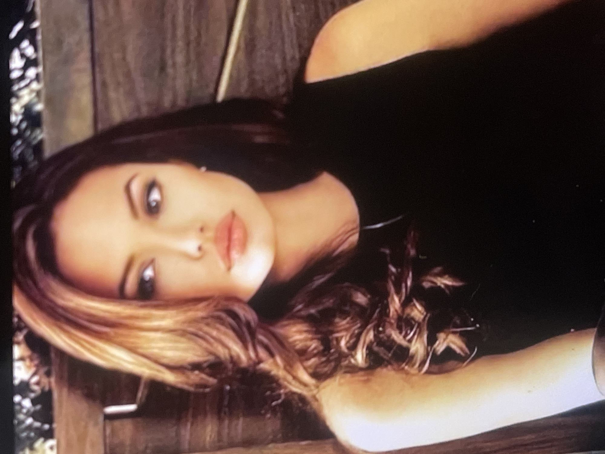 A striking portrait features a woman, resembling actress Angelina Jolie, lying down and looking directly into the camera. Her head is positioned on the left side of the frame, tilted slightly, with her gaze fixed and intense. She has long, wavy hair, dark at the roots and transitioning to lighter, highlighted strands, which spreads out around her head. Her characteristic full lips are subtly parted, and her eyes are dark and captivating. She wears a simple, dark, possibly black, sleeveless garment.
The woman is reposed on a surface composed of broad wooden planks, suggesting a bench, deck, or similar structure. To the left of her head, the background shows blurred foliage or greenery, indicating an outdoor or semi-outdoor setting. The lighting softly illuminates her face, emphasizing her features and adding a warm glow to her skin. The image conveys a mood of confident allure. There is no visible text present in the media file. While the location context is Poltava, Ukraine, the visual details within the image do not provide specific cues to confirm this geographical location.