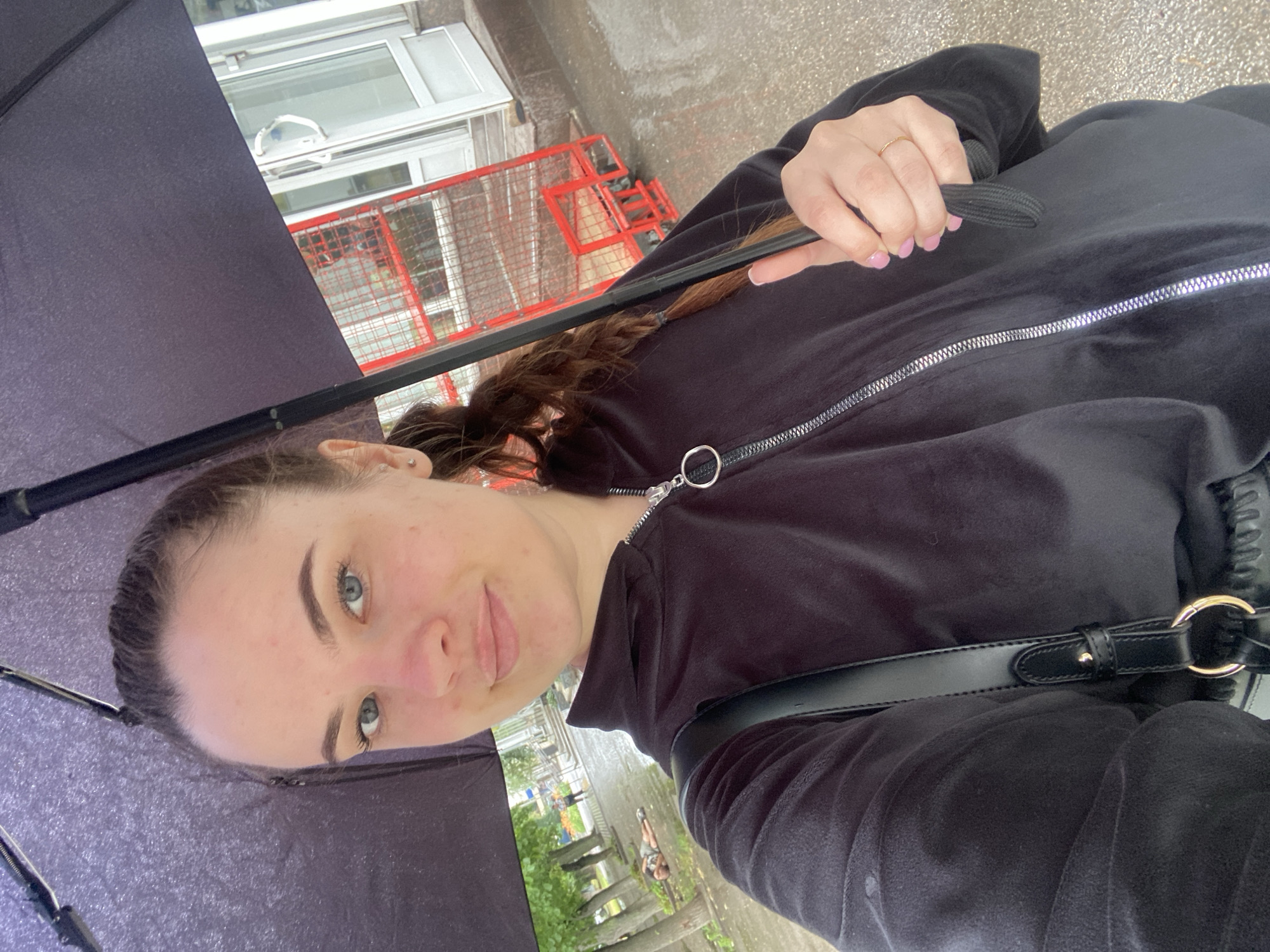 A young woman with light skin and light blue eyes is taking a selfie outdoors while holding a dark umbrella. She has brown hair pulled back in a ponytail, and she is wearing a black hooded jacket with a silver zipper. A black purse strap is visible over her left shoulder. Her right hand is holding the umbrella handle, and she is wearing a thin gold ring on her right ring finger.
The background suggests an urban environment. To the right of the woman, there is a building with white doors and windows, and a red metal structure that appears to be a trolley or cart. To the left and behind her, there are trees lining a wet, paved area, possibly a sidewalk or street, indicating it might be raining or has recently rained. In the distance, a person can be seen walking, and another person is lying down on the ground near the trees. The overall scene appears to be a street in a town or city. The location is identified as Nyzhni Mlyny, Ukraine.