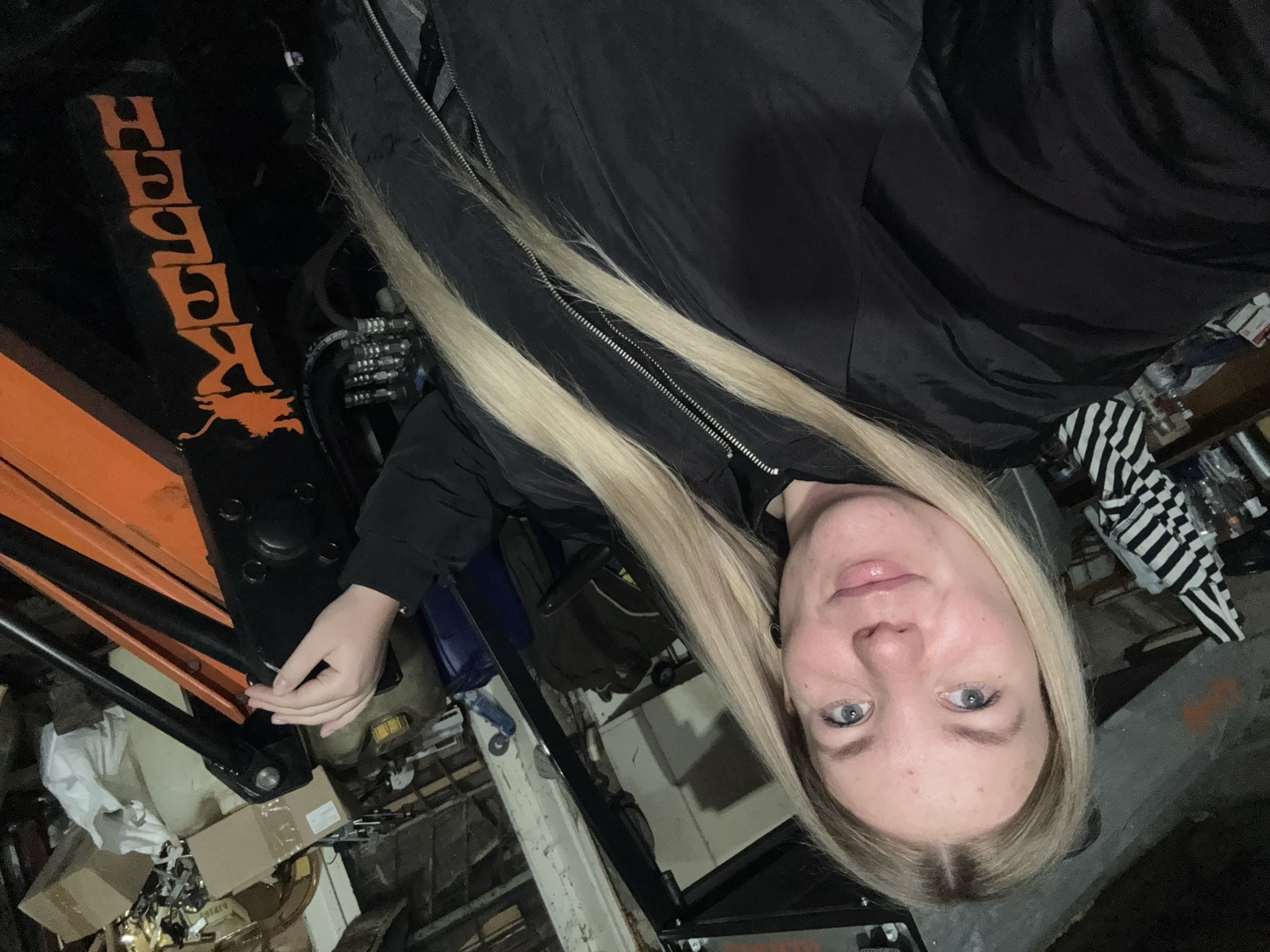 A person with long blonde hair, wearing a black jacket, is partially visible in a cluttered indoor location. Adjacent to the person is an orange and black machine with the word "KALEKH" printed vertically on its black surface. The surroundings suggest a workshop or storage area filled with various items, including boxes, containers, and other mechanical parts. The specific location in the city of Chaikovsky, Russia, is not discernible from the image.