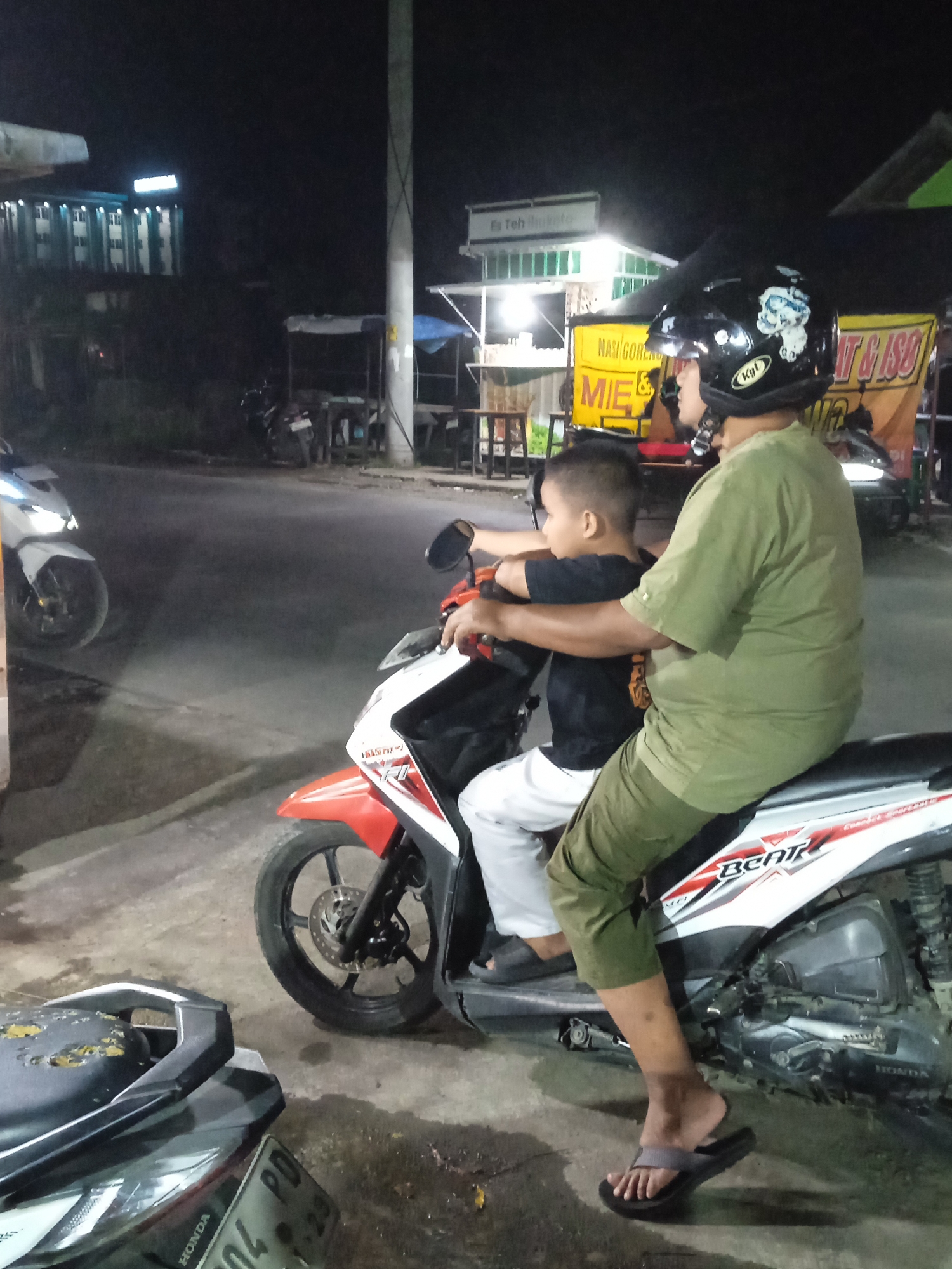The image captures a night scene in Kebonsari, Indonesia, showing an adult and a child on a red and white Honda Beat scooter. The adult, wearing an olive green shirt and pants with flip-flops, is in the driver's position, donning a black full-face helmet with a "KYT" logo and a white cartoon sticker. A young boy, dressed in a black t-shirt and white pants, sits in front of the adult, holding the handlebars. Both are looking forward, suggesting they are either stopped, waiting, or just beginning a journey.

The setting is a street illuminated by artificial lights, indicating it is nighttime. In the background, various roadside stalls are visible with bright lights and banners. One banner partially reads "NASI GORENG & MIE," advertising food, and another mentions "Es Teh." A multi-story building with lighted windows can be seen further back. Another white scooter is partially visible on the left side of the road, and a dark scooter is in the immediate foreground, with a partially visible license plate starting with "PD." The overall impression is a typical bustling night scene in an Indonesian urban or semi-urban area.