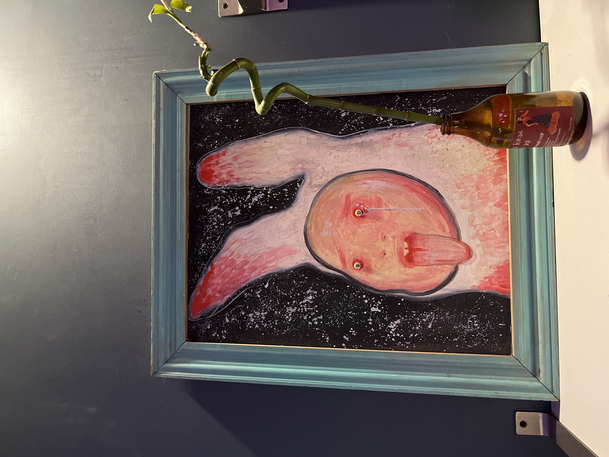 The image depicts a painting in a light blue frame with a distressed finish, hanging on a dark gray wall. The painting itself is an abstract or surreal portrait of a creature with large, floppy pink ears or hands, a round, pale face with wide, bloodshot eyes, and a protruding tongue. The background of the painting is black with white speckles. To the right of the painting, a twisted green bamboo stalk in a brown glass bottle stands on a white surface. The bottle has a label with a graphic of a woman in a dress. The overall scene suggests an indoor setting, possibly a home or a studio, and does not provide any specific visual cues to pinpoint a location within Saint Petersburg, Russia.