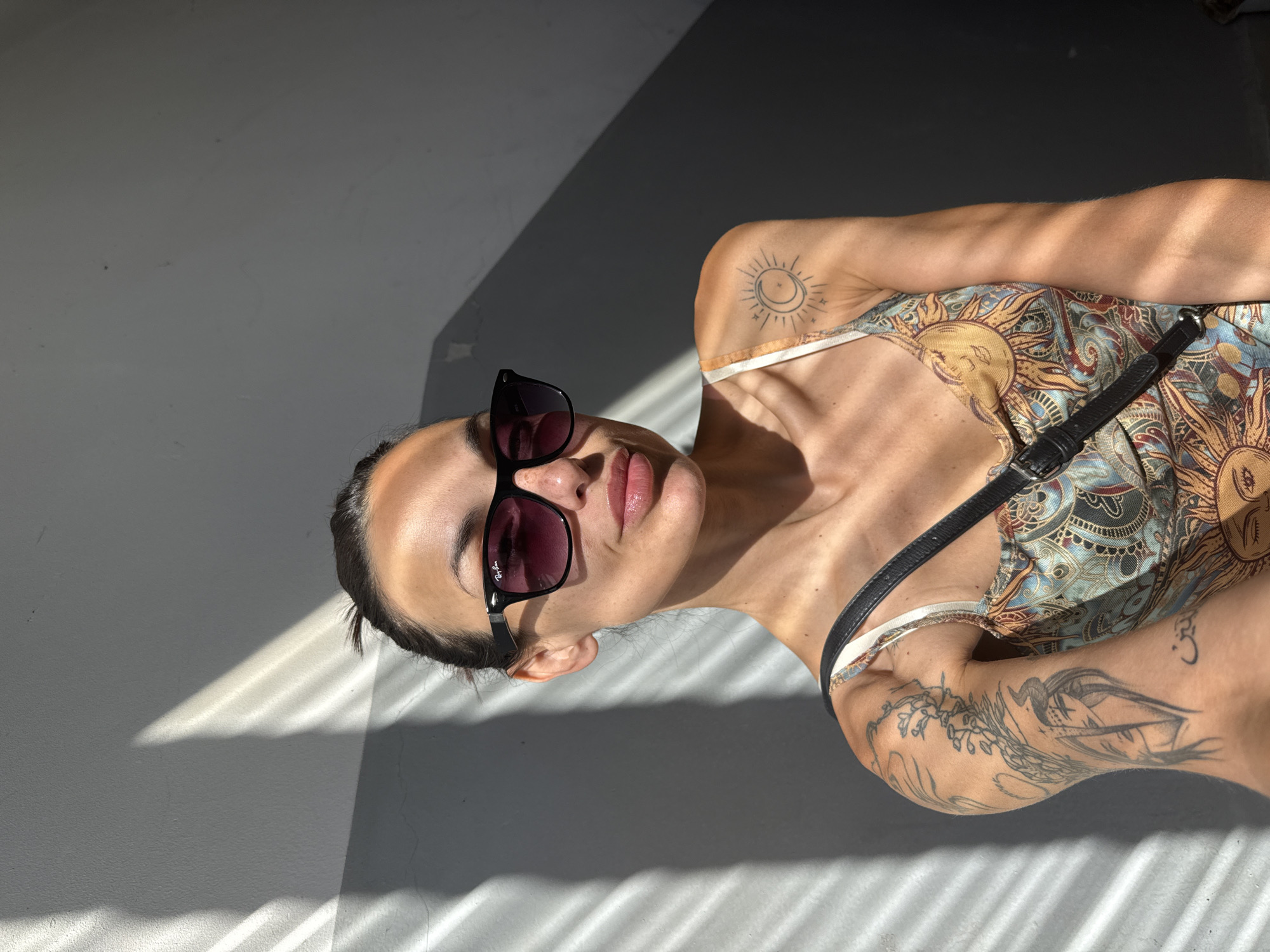 A woman with dark hair pulled back in a bun, wearing sunglasses and a patterned dress with sun motifs, poses for a selfie against a grey, textured wall. Sunlight casts diagonal stripes of light and shadow across the wall and the woman's face and left arm. She has visible tattoos on her left arm, including a design featuring a woman's face with horns and what appears to be floral elements. A small tattoo of a sun and crescent moon is visible on her right shoulder. The scene is outdoors, with the harsh sunlight suggesting a bright day. The setting is not specified to be in the city of Pleven, Bulgaria.