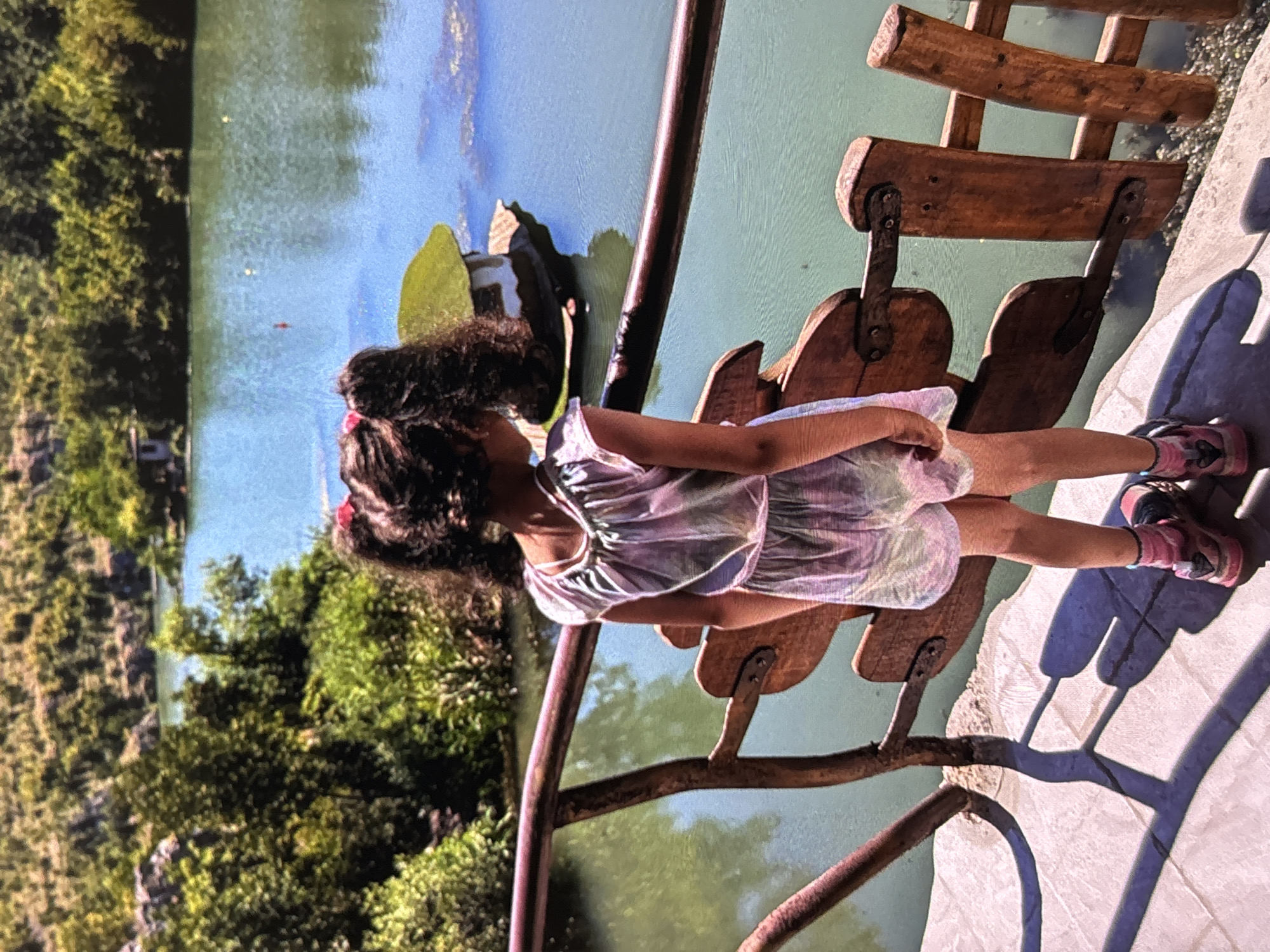 The image captures a young girl standing beside a rustic wooden bench or swing, gazing out over a tranquil body of water. The girl has dark, curly hair styled with hair ties and is dressed in a shimmering, light-colored dress or top. She wears pink and black athletic shoes with striped socks. Her posture suggests a moment of quiet observation as she looks away from the viewer towards the expanse of water.
The wooden structure she is next to features curved slatted seats and a dark, possibly metal, frame or railing. This structure is positioned on a paved or concrete area. Long, distinct shadows are cast across the ground, indicating a bright, sunny day, likely midday or early afternoon. The water, which appears to be a lake or large pond, shows reflections of the blue sky and surrounding green foliage, suggesting a clear sky.
The background is dominated by lush, green trees and dense vegetation that line the distant banks of the water body, creating a natural and verdant setting. The scene evokes a peaceful outdoor environment, possibly a park, nature reserve, or a scenic overlook. No visible text is present in the image.