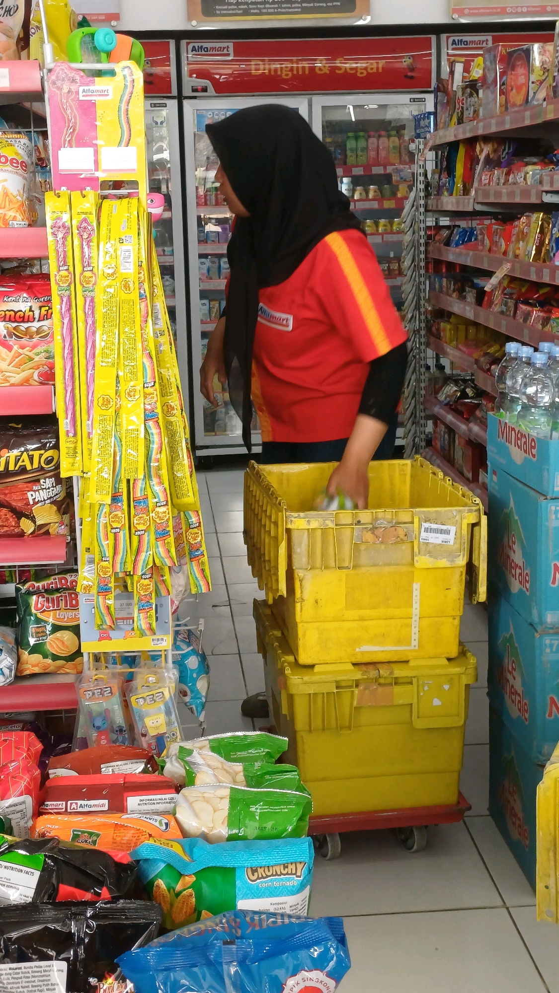 The image shows a woman in a red Alfamart uniform, wearing a black hijab, in what appears to be an Alfamart convenience store. She is standing next to shelves stocked with various snacks and drinks. In front of her are two yellow plastic bins stacked on a red dolly, and she is in the process of placing items into the upper bin. The floor is tiled, and there are aisles of products visible in the background, including refrigerators displaying drinks. The presence of "Alfamart" branding throughout the store indicates it is a chain of convenience stores common in Indonesia. The scene suggests a retail environment where inventory management or restocking is occurring. Based on the Alfamart branding, this location is likely in Tengahan, Indonesia, as Alfamart is a prevalent store chain there.