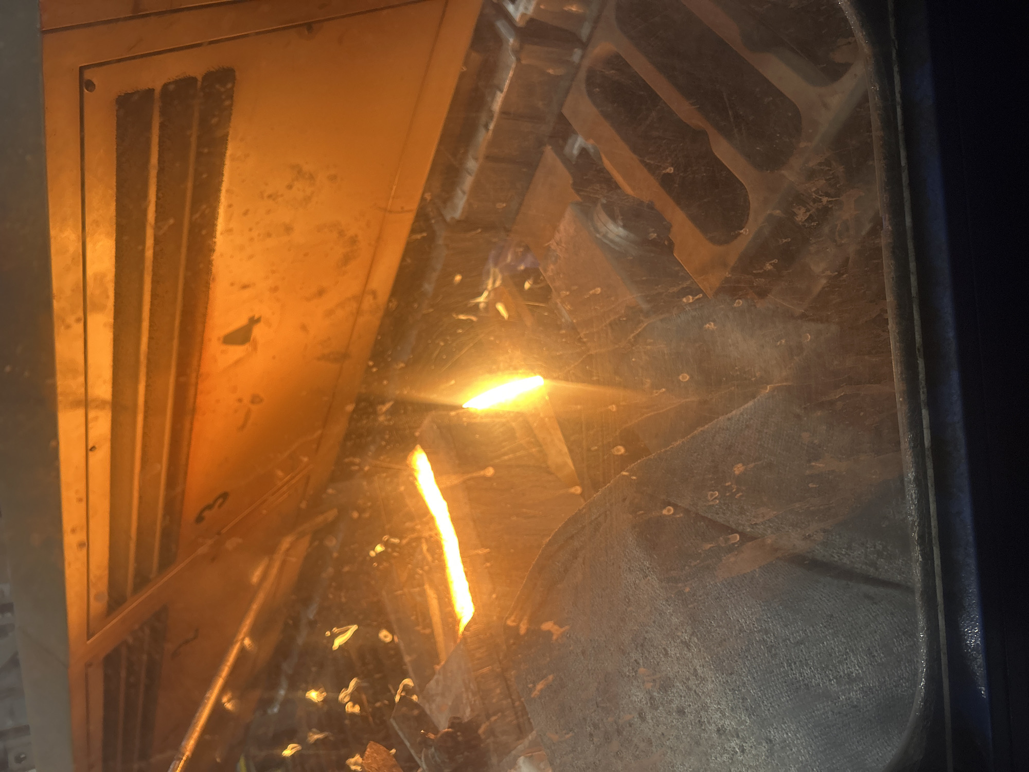 The image depicts the inside of a fireplace, likely a wood-burning stove, with flames visible. The internal components of the fireplace, including metal grates and structures, are visible through a dirty glass door. The scene is interior, illuminated by the fire. No people are visible. There is no information regarding location.