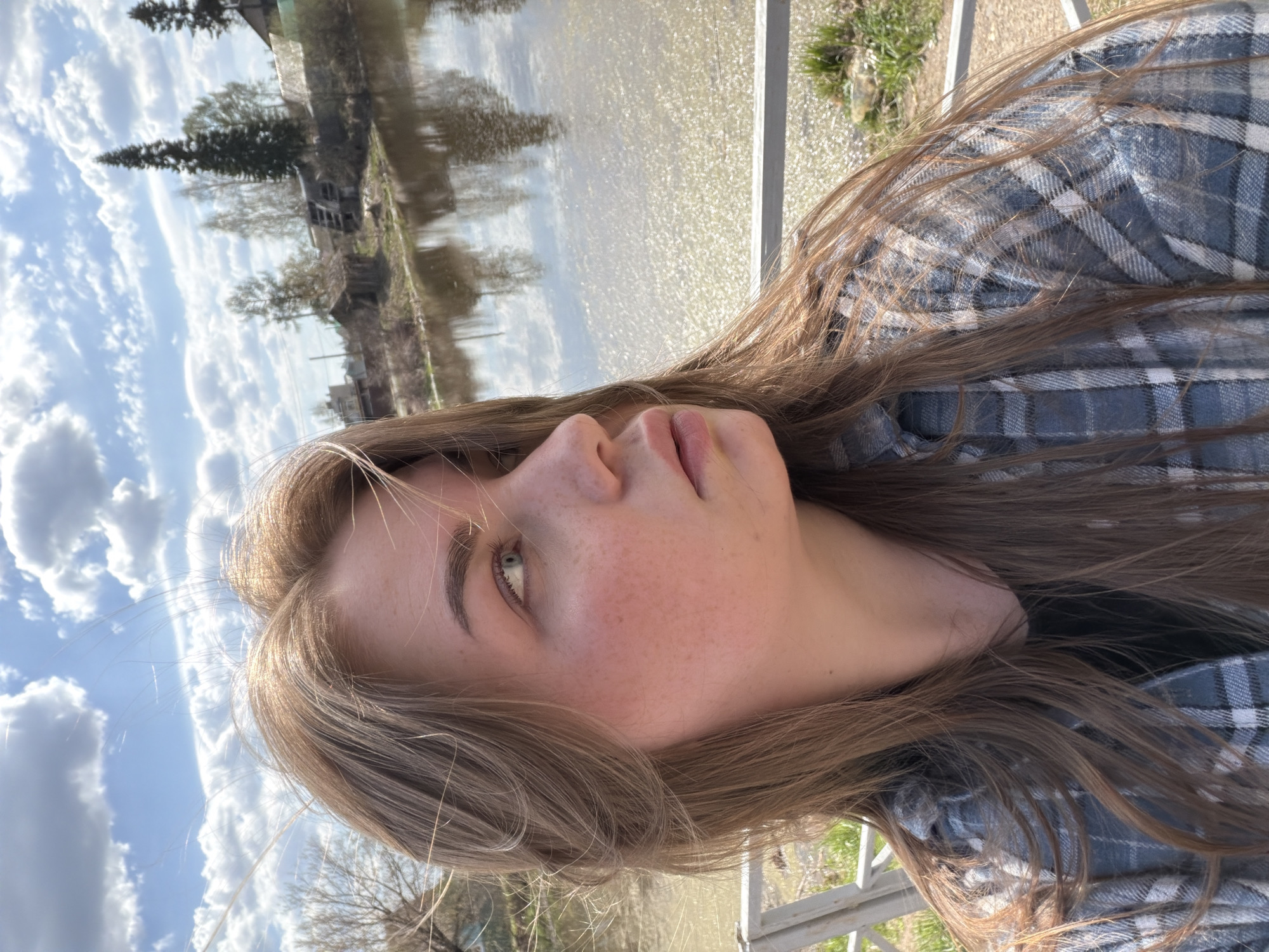 A medium shot shows a young woman in a blue plaid shirt, with light brown hair and fair skin. She is standing on a bridge over water reflecting the sky and buildings. The location is speculated to be in the city of Chaikovsky, Russia, based on potential identifying factors in the reflection. The woman is looking off into the distance. The sky is partly cloudy. A metal railing is visible in the foreground.