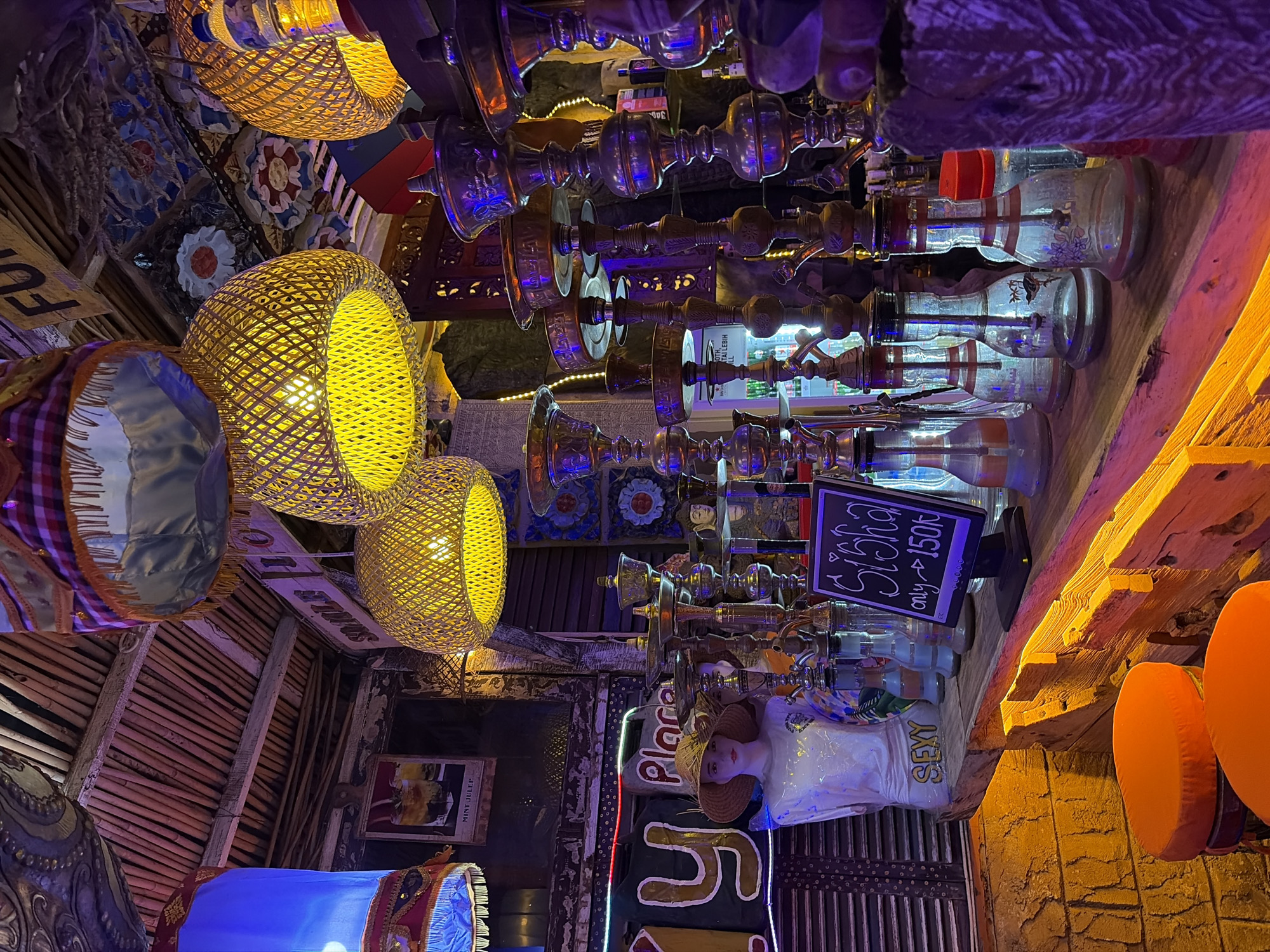 This is a medium-shot, eye-level interior photograph of a hookah lounge or bar. The scene is dimly lit, with warm yellow and purple lighting creating an ambient atmosphere. The foreground features a wooden bar counter with several ornate brass hookahs arranged on it. The hookahs have glass bases and intricate metal stems. To the left of the hookahs, there is a mannequin dressed in a white shirt and a straw hat, with the word "SEXY" printed below it. Behind the mannequin, a neon sign spelling "PLAY" is visible. Further in the background, there are shelves with various items, including what appear to be bottles. Hanging from the ceiling are multiple woven lampshades emitting a warm yellow glow, and other decorative elements. The ceiling is constructed of bamboo or similar material. A small blackboard sign on the bar displays "Sisha only - 150k". Two orange stools are visible at the very bottom left of the image. The overall aesthetic suggests a relaxed and perhaps exotic setting.