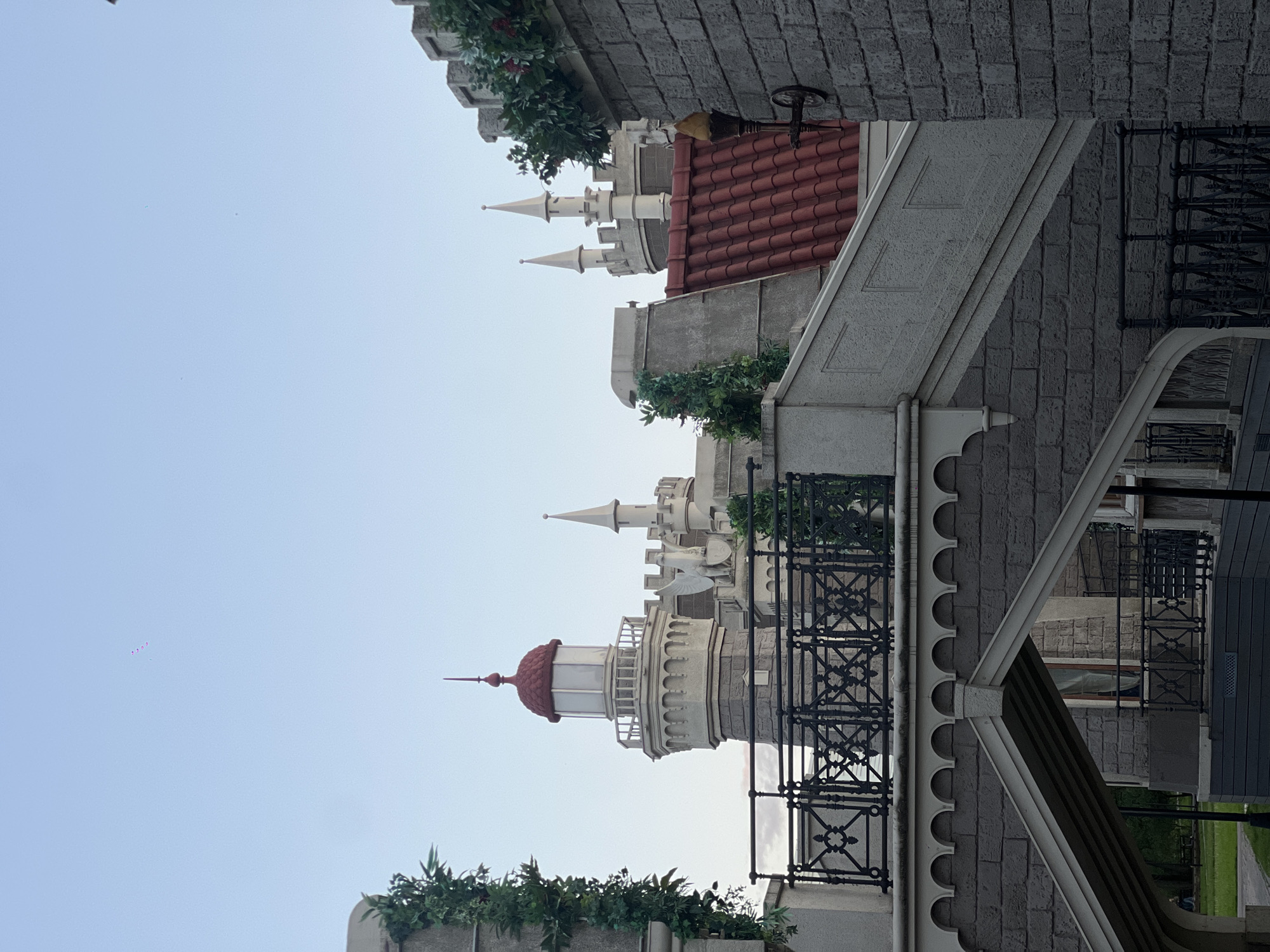 The image shows a building complex in Moscow, Russia, with a fanciful architectural style reminiscent of a fairytale castle. The buildings are constructed from light gray stone and feature decorative elements such as towers with pointed spires, red-tiled roofs, and ornate balconies with black metal railings. There are plants and greenery on top of the buildings. The sky is pale blue.