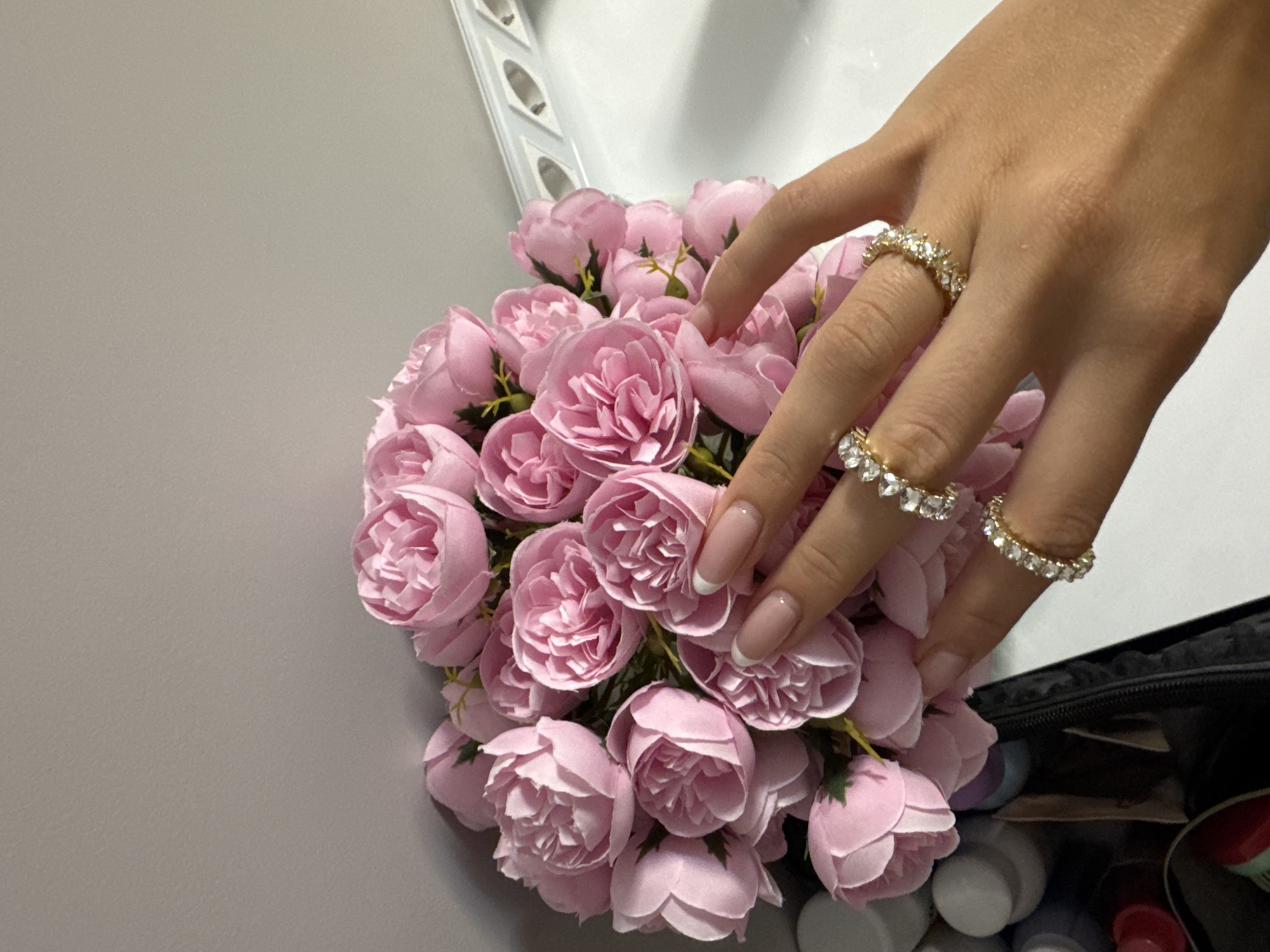 A hand with three rings (a diamond band on the ring finger, a diamond band on the middle finger, and a diamond band on the index finger) rests atop a bouquet of pink roses. The hand has fingernails with a white French manicure. The bouquet is set against a grey surface on the left and a white surface with a white object on the top left. On the bottom right, there is a black object containing various white, black, and red items. The location cannot be determined.