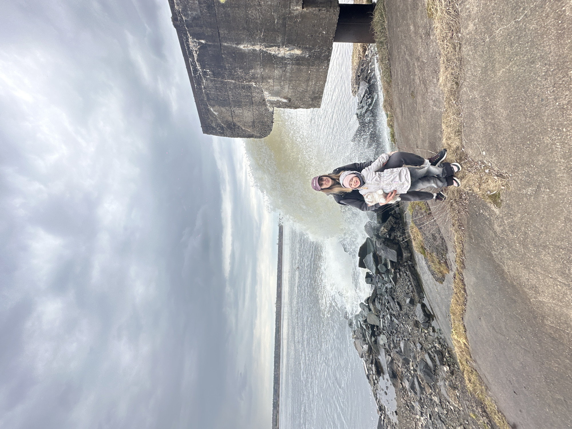 A mother and child stand next to a body of water, likely a river or reservoir, next to what appears to be a concrete structure with water flowing from it. The location is Chaikovsky, Russia, as indicated by the image context. The mother wears a dark jacket and a purple hat, and she holds a young child who is wearing a light-colored jacket. The environment is overcast with a gray sky. A concrete pathway is present next to the water and the concrete structure. Rocks are present at the water's edge, and the water itself appears turbulent.