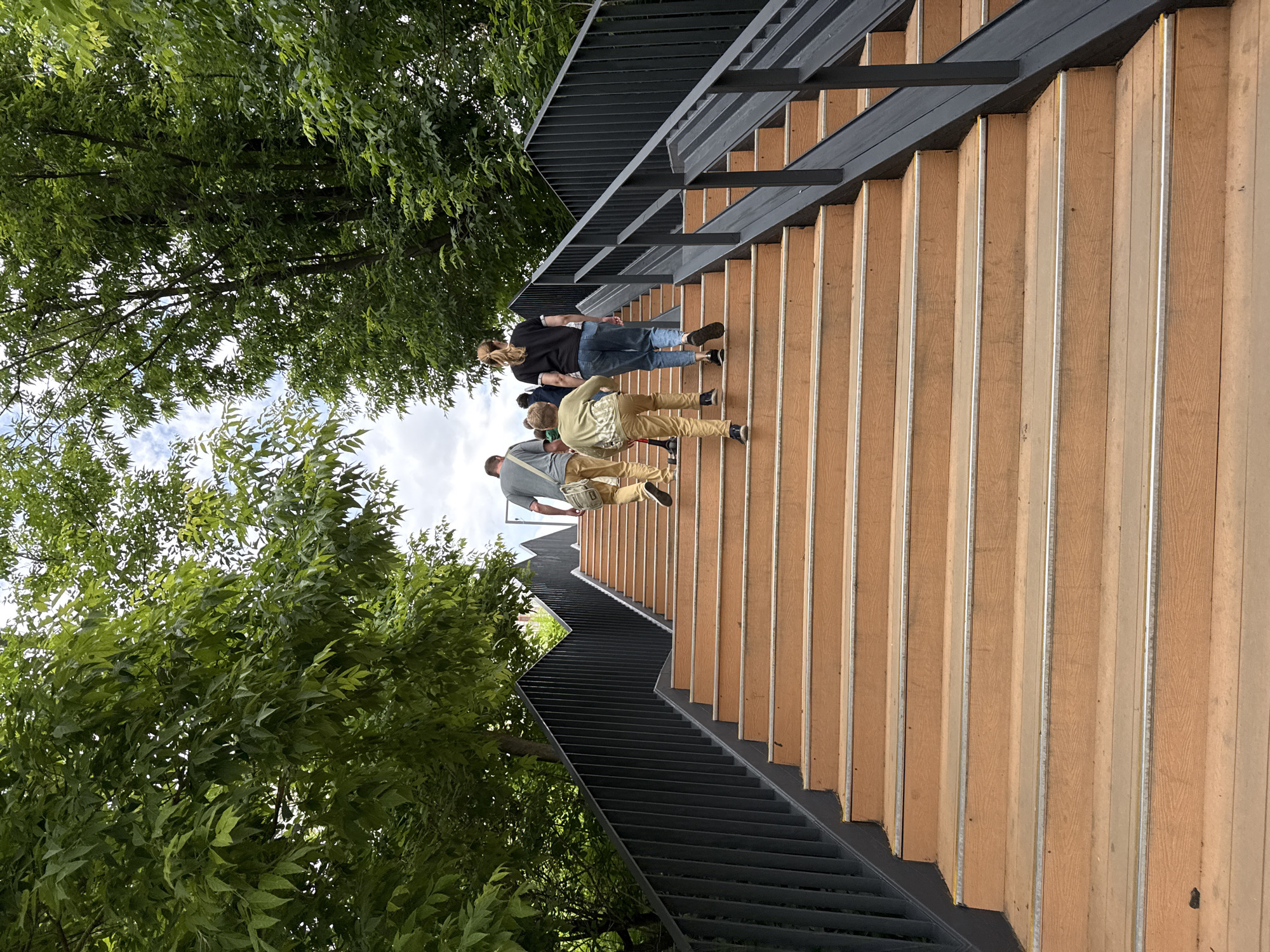 In Moscow, Russia, a group of four individuals, two adults and two children, ascend a wide, outdoor wooden staircase with black metal railings. The man in front wears a gray t-shirt and light brown pants, carrying a beige messenger bag. Behind him, a young boy in a light green long-sleeved shirt and tan pants walks with a scooter tucked under his arm. To his right, a woman in a black t-shirt and blue jeans walks ahead of a smaller child. The staircase is flanked by lush green trees, their leaves creating a canopy overhead, and the sky is overcast with white clouds. The wooden steps have metal edging, and the railings are made of vertical black metal bars. The scene appears to be in a park or public outdoor area, with the staircase possibly leading to an elevated viewpoint or connecting different levels of the park.