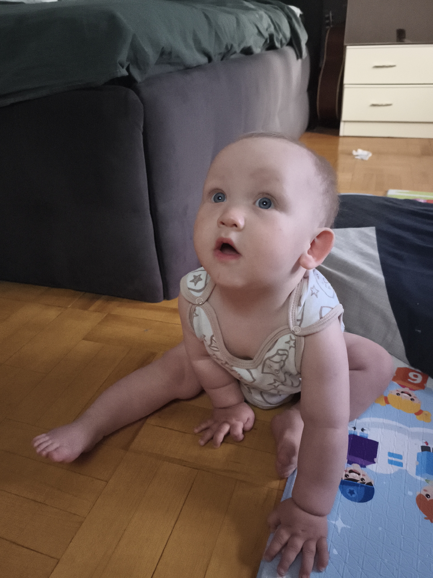 The image shows a baby with blue eyes and light hair sitting on a wooden floor. The baby is wearing a light-colored onesie with a star pattern. The baby's legs are spread apart, and its arms are resting on a blue playmat with cartoon characters. In the background, there is a dark gray sofa, a green blanket, and a chest of drawers. The overall scene appears to be in a residential setting. There is no visible indication of the city of Chaikovsky, Russia.