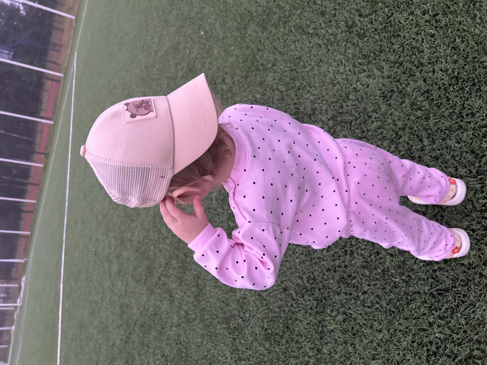 A young child, viewed from behind, stands on a green artificial turf field, likely a sports field, in Noviy, Russia. The child is wearing a pink, polka-dotted long-sleeved shirt and pants, and white sneakers with floral accents. On their head, the child wears a light pink baseball cap with a mesh back and a patch depicting a cartoon bear. The child's left hand is raised to their ear, as if adjusting the cap or listening. The field has white markings visible, and in the background, tall poles and trees suggest an outdoor, possibly park or recreational area. The lighting indicates it may be overcast or late in the day.