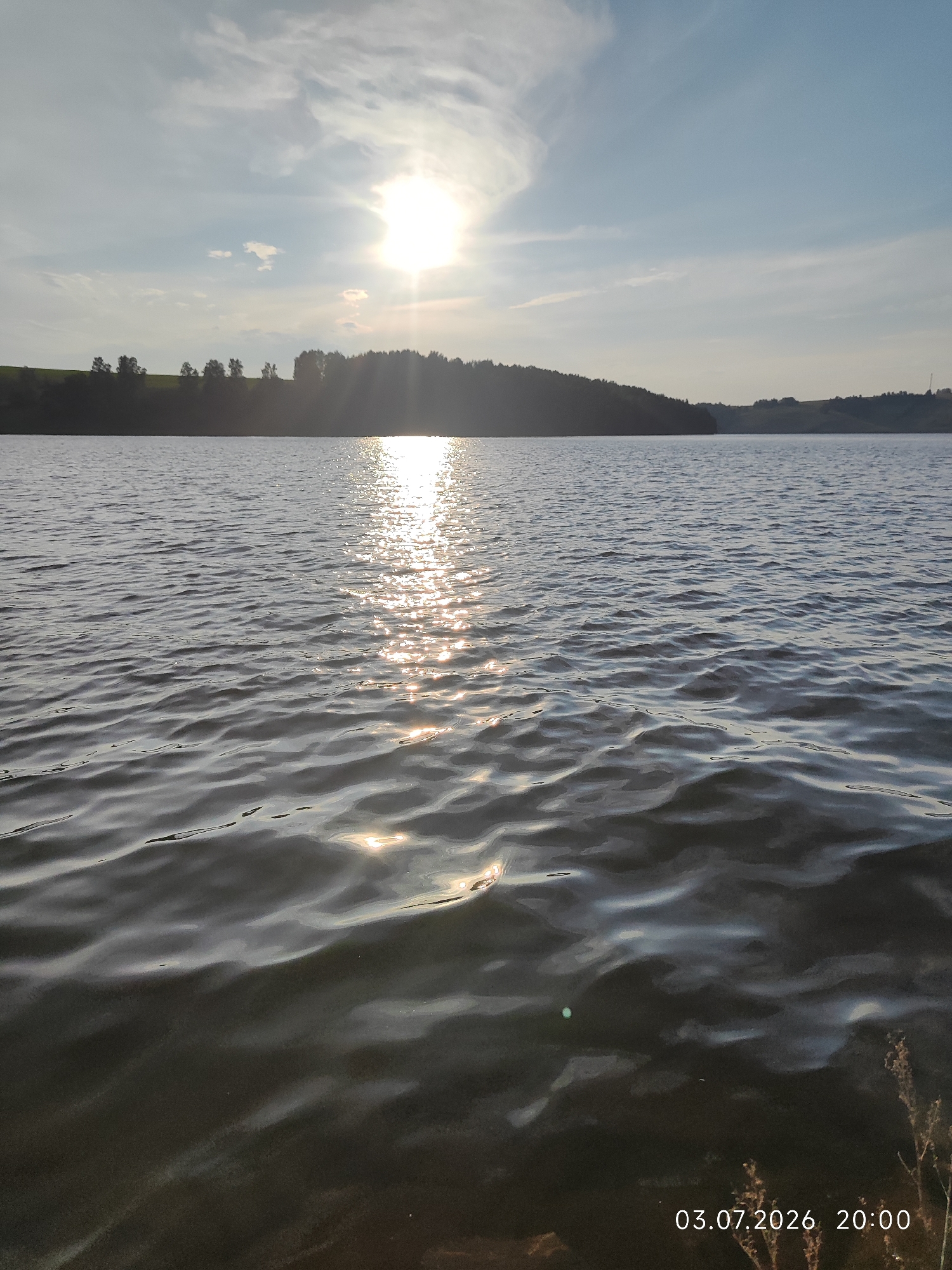 The image depicts a tranquil sunset over a body of water, likely a lake or reservoir, in Konovalovo, Russia. The sun, a brilliant orb, is positioned in the upper center of the frame, casting a shimmering path of light across the gently rippling surface of the water. The water itself is a deep, dark blue, reflecting the sky and the sun's rays.
In the background, a silhouetted treeline forms a natural horizon, punctuated by a gently sloping hill dotted with more trees. The sky is a canvas of soft blues and whites, with wispy clouds catching the last golden light of the day. The overall atmosphere is serene and peaceful, evoking a sense of calm and natural beauty.
The time of day is clearly indicated by the date and time stamp in the lower right corner: 03.07.2026 20:00. This suggests it is early evening in the summer. The weather appears to be clear and pleasant, with the sun still strong enough to create a vivid reflection. There are no people or man-made objects prominently featured in the scene, emphasizing the unspoiled natural landscape. The focus is entirely on the interplay of light, water, and the surrounding environment. The image captures a moment of quiet contemplation, inviting the viewer to appreciate the simple elegance of a sunset by the water.