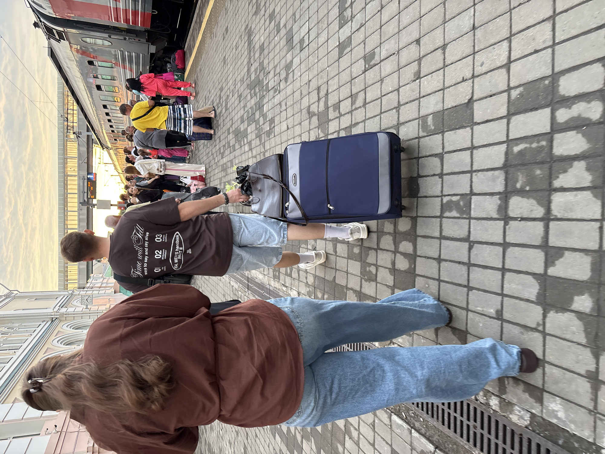 The image shows a train station platform in Khabarovsk, Russia, with several people waiting for or disembarking from a train. The sky is overcast with hints of a sunset. The platform is paved with grey bricks, and a train with red and grey carriages is visible on the right side of the image. In the foreground, two individuals are walking away from the camera. The person on the left is wearing a brown oversized shirt and light blue flared jeans, with their long brown hair tied back in a ponytail. The person on the right is wearing a dark brown t-shirt with text and graphics, light blue denim shorts, and white sneakers with grey socks. They are pulling a dark blue wheeled suitcase topped with a brown leather bag. Other passengers are visible in the middle ground, some with luggage, suggesting travel. The architecture of the station building is partially visible in the background, featuring arched windows.