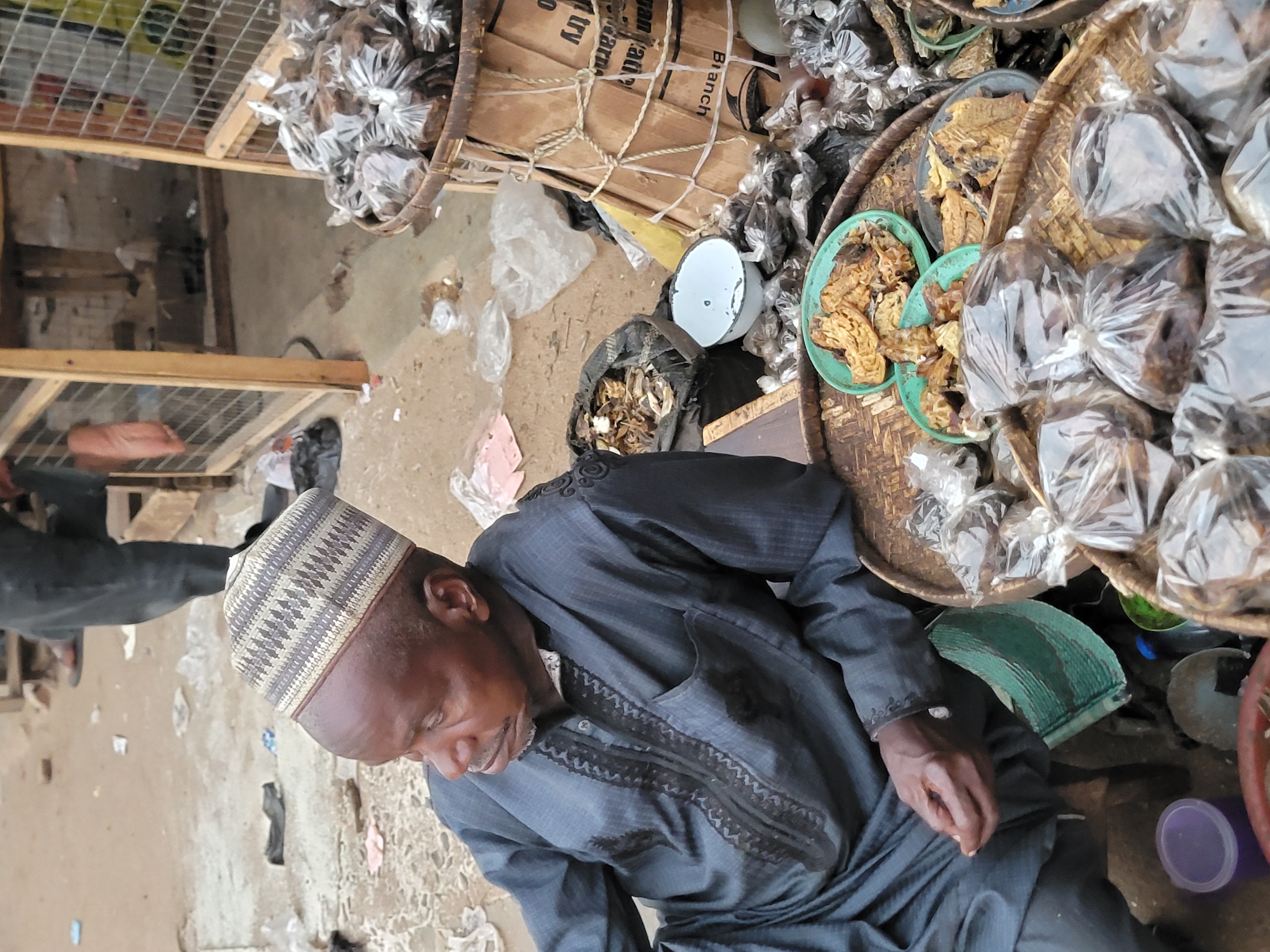 A man wearing a patterned cap and a dark blue embroidered shirt is seated at an outdoor market stall. To his right are several woven baskets filled with items, including those packaged in clear plastic bags. To the left is a bucket with wooden boards secured by rope. Other various items are scattered around the stall. The scene appears to be in a market in Jos, Nigeria.