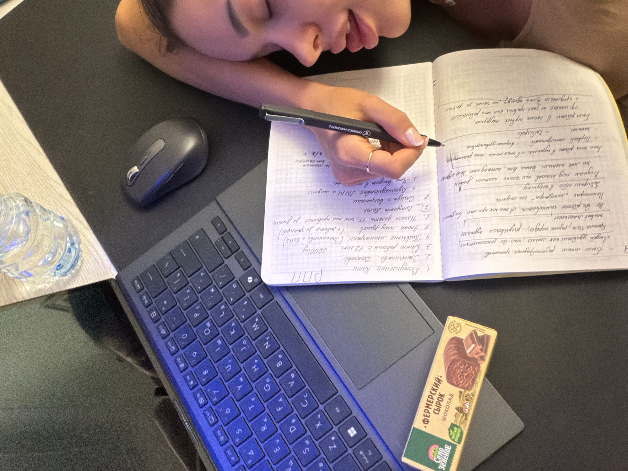 An overhead shot captures a person, likely a woman, resting her head on her arm while writing in a notebook at a desk. Her eyes are closed or mostly closed, suggesting deep concentration, relaxation, or fatigue.
The scene, set indoors in what appears to be Moscow, Russia, depicts a workspace with several items arranged on a dark surface, possibly a large mouse pad or desk mat. To the left, a partially visible plastic water bottle sits next to a dark grey computer mouse. A laptop with a black keyboard, featuring Cyrillic characters illuminated with a blue hue, is positioned in the lower left and center of the frame.
The person holds a black pen, branded "TURKISH CARGO," as she writes in an open notebook filled with handwritten Cyrillic text. The writing includes lists and paragraphs, with discernible phrases such as "Вступление Кати" (Introduction of Katya), "Знакомство. Детство" (Acquaintance. Childhood), "Опыт работы с 12 лет" (Work experience from 12 years old), and "Взаимодействие SMM и модели" (Interaction between SMM and model). Other notable phrases include "Было много разнообразных проектов" (There were many diverse projects) and mentions of "инстаграм" (Instagram) and "2006reg."
In the lower right corner, a rectangular package for a curd snack, bearing the brand "Село Зеленое" (Selo Zelenoe - Green Village) and labeled "ФЕРМЕРСКИЙ СЫРОК ШОКОЛАД" (Farmer's Curd Snack Chocolate) with "чистый продукт" (pure product) visible, rests on the laptop's palm rest. The overall impression is of a quiet moment of study or work.