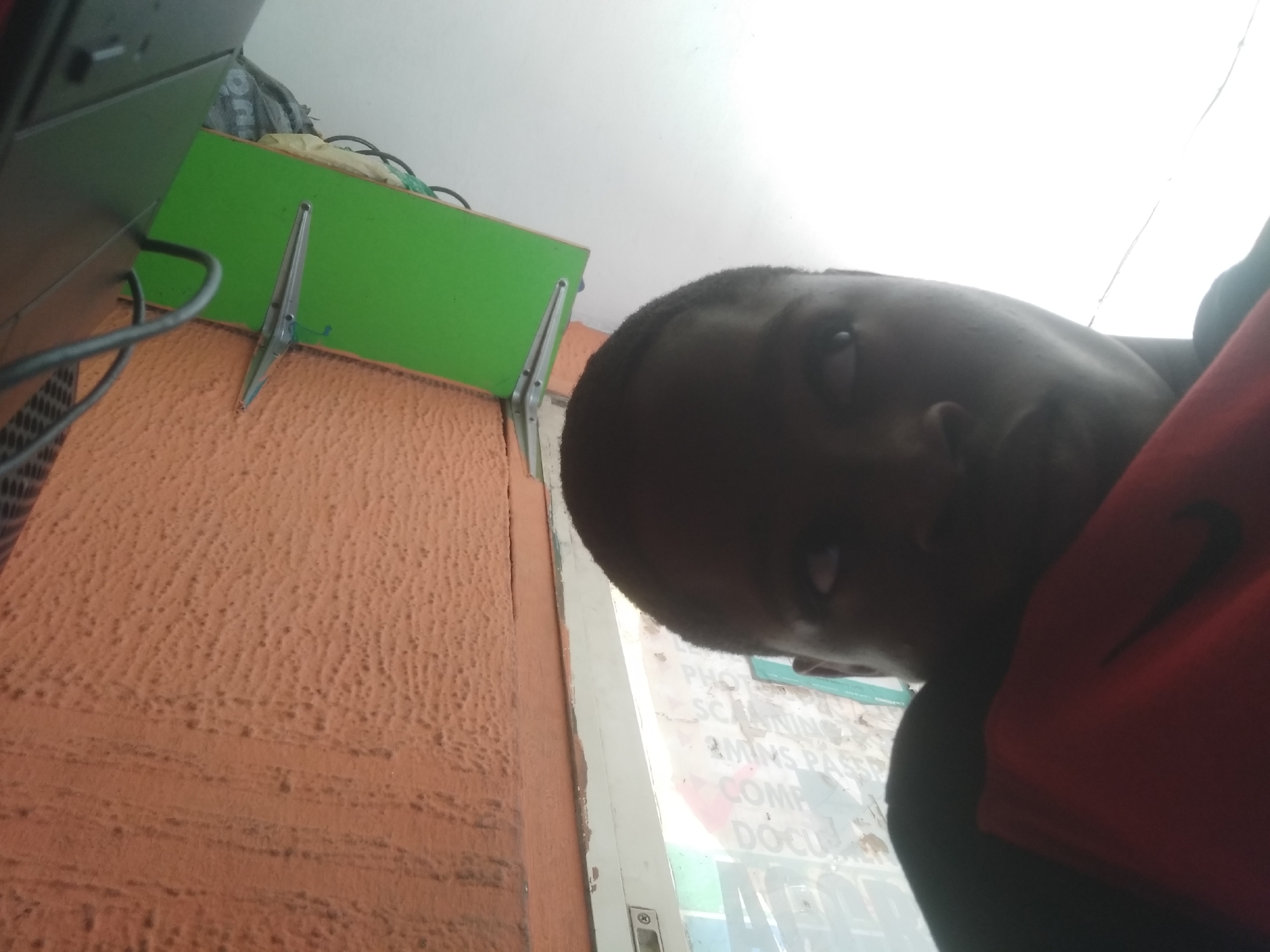 A young individual, likely male, with dark skin and short dark hair, is captured in a close-up, angled selfie. Their face is partially shadowed, but one eye is clearly visible, looking directly at the camera. They are wearing a red top, possibly a t-shirt or hoodie, with a black logo resembling the Nike swoosh on the right side of their chest.
The setting appears to be an indoor or sheltered space. To the left, a textured, rough orange-brown wall occupies a significant portion of the frame. Above it, a green shelf or surface is visible, connected to the wall by a metal bracket. Wires and cables, some black, are draped over the shelf and an adjacent dark electronic device, possibly a computer monitor, with a perforated side.
To the right, behind the person, there's a white door or window frame, with a latch or lock mechanism visible at the bottom. Through the glass, bright daylight suggests it's daytime, and several words are reflected or visible, indicating a business-related environment. The discernible text includes "PHOTO," "SCANNING," "3 MINS PASS" (likely "3 Minutes Passport"), "COMP" (possibly "COMPUTER"), and "DOCU" (likely "DOCUMENTS"), suggesting a place offering photography, scanning, passport, and document services.
The perspective is tilted, consistent with a handheld selfie taken from an unusual angle, possibly looking up at the phone. The overall impression is that of a casual moment captured in a small business or utility area within Shangisha, Nigeria.