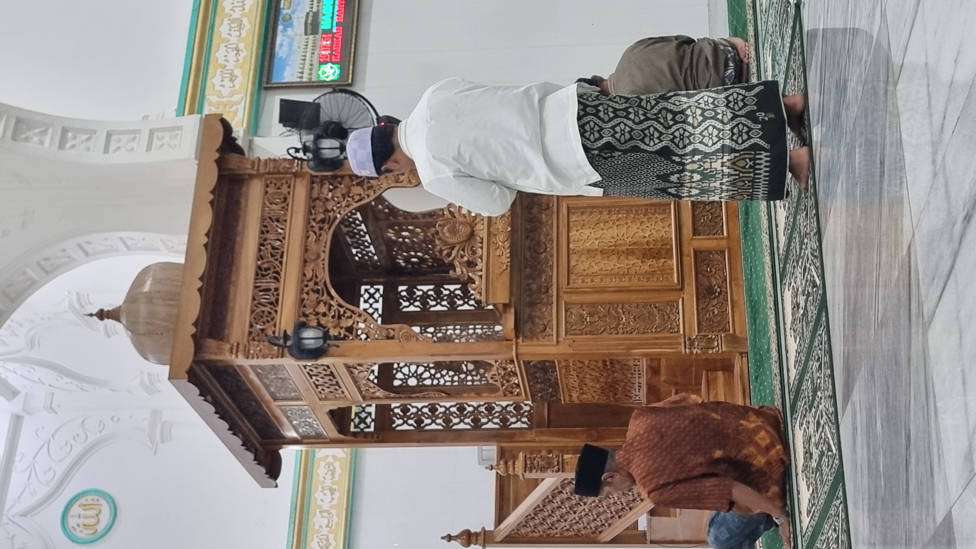 This is a photograph taken inside a mosque in Lapang Timu, Indonesia. The central focus of the image is a beautifully carved wooden minbar, a raised platform from which sermons are delivered, positioned in front of a large arched opening. To the left of the minbar, a person is seated on the floor, facing away from the camera, wearing a traditional brown patterned shirt and a black skullcap. To the right of the minbar, another individual stands with their back to the camera, wearing a white shirt and a patterned sarong, with a white cap on their head. This person appears to be facing the minbar. In the background, a digital screen displays time and text in a language likely Indonesian. The floor is covered with green prayer mats and a polished marble-like material. The mosque's interior architecture includes white arched ceilings and decorative elements. The overall scene conveys a sense of religious observance and architectural craftsmanship.