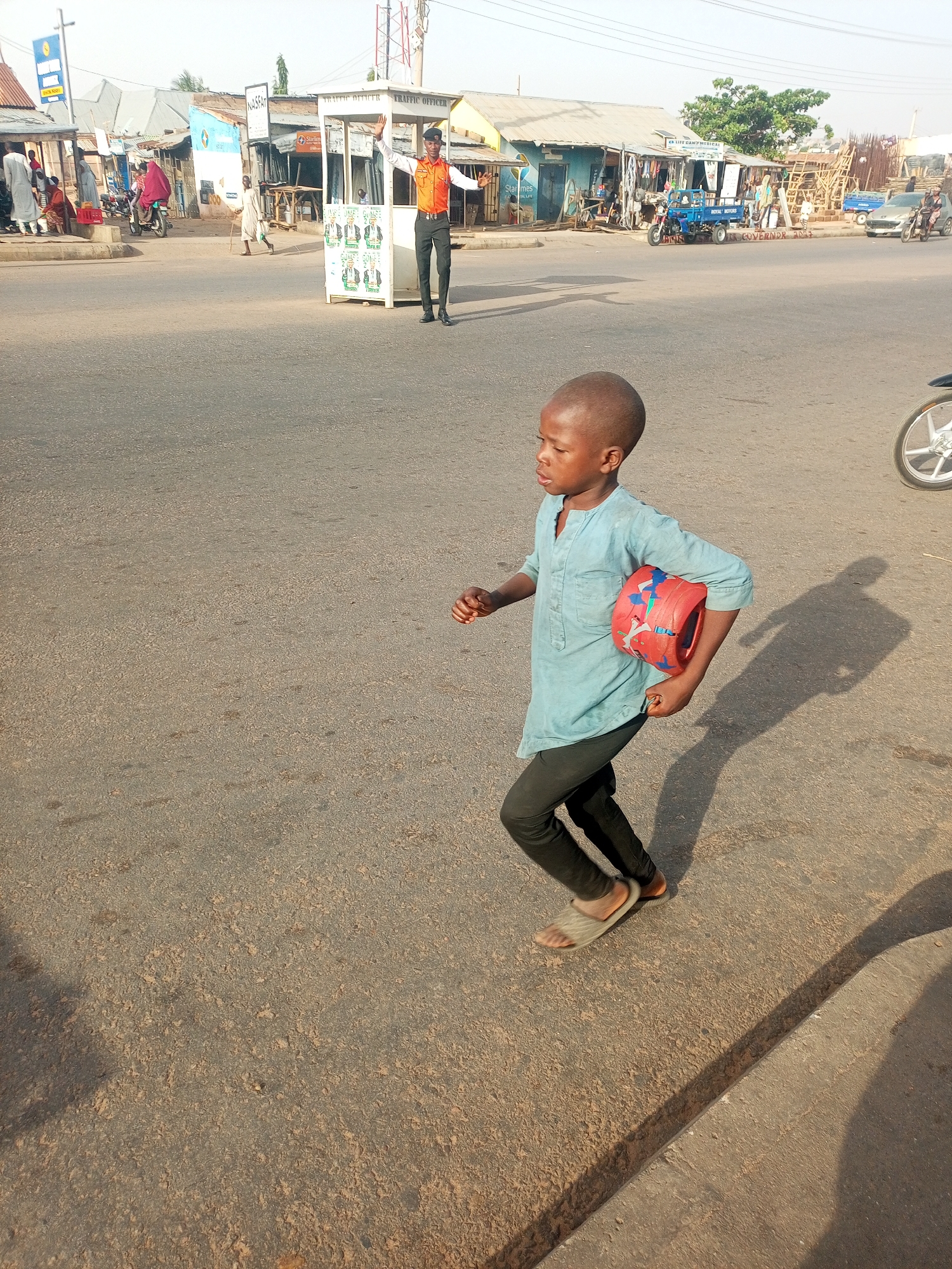 A young boy, dressed in a light blue shirt and dark pants, is running across a paved road. He's holding a red and blue cylindrical object. His shadow is cast on the road next to him. In the background, a traffic officer stands near a small white booth, directing traffic with his arms outstretched. There are several buildings and shops lining the road. Vehicles, including motorcycles and a car, are visible in the background. The location is Gombe, Nigeria, based on the "Gombe state" electioneering on the traffic officers' booth.