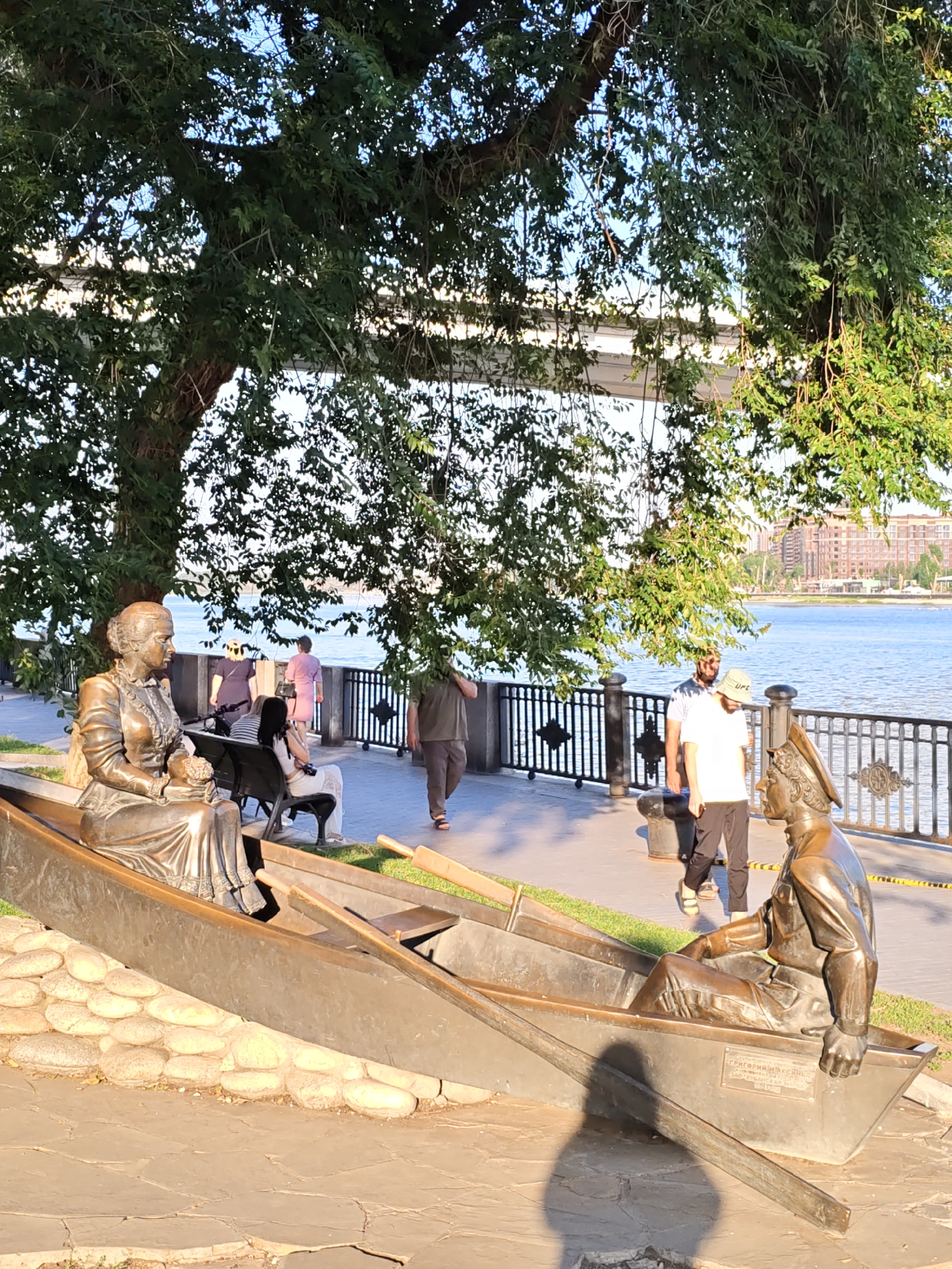 The image depicts a bronze sculpture of two figures in a boat, situated on a paved promenade overlooking a body of water. The sculpture is located at the Rostov-on-Don embankment, a popular waterfront area in the city of Rostov-on-Don, Russia. The sculpture itself features a woman in a long dress seated on the left, holding a bouquet of flowers, and a man in a military-style uniform seated on the right, looking forward. They are positioned within a bronze boat that rests on a stone base.
In the background, several people are walking along the promenade. To the left, a person is seated on a bench. A metal railing separates the promenade from the water. Beyond the railing and the water, buildings can be seen on the opposite bank, indicating an urban environment. Large trees with lush green foliage extend over the promenade, casting shadows and providing shade. The sky above is blue with some sunlight filtering through the leaves. The overall scene suggests a public space, likely a tourist attraction or a recreational area.