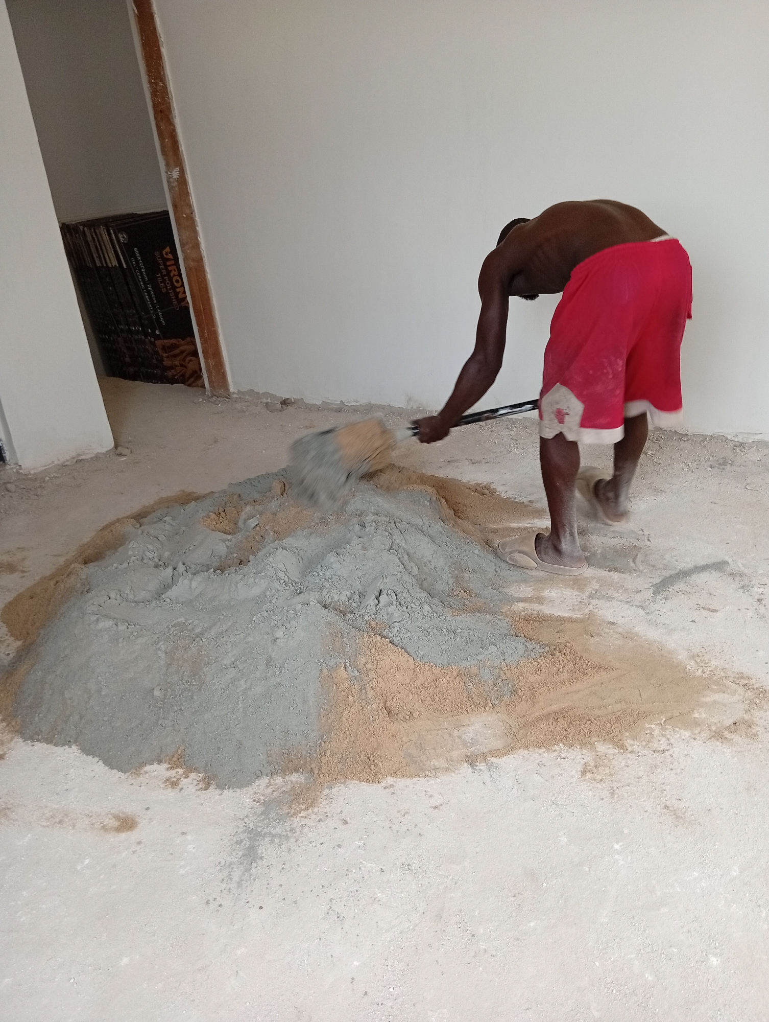 The image depicts a dark-skinned man in red shorts mixing a pile of gray and tan substances, likely cement and sand, on a light-colored, possibly concrete, floor inside a building. He uses a long-handled tool to combine the materials. In the background, stacks of dark-colored tiles with the brand name "VIRONY" are visible near a doorway. The location appears to be a construction site in a city of Unknown, Unknown.