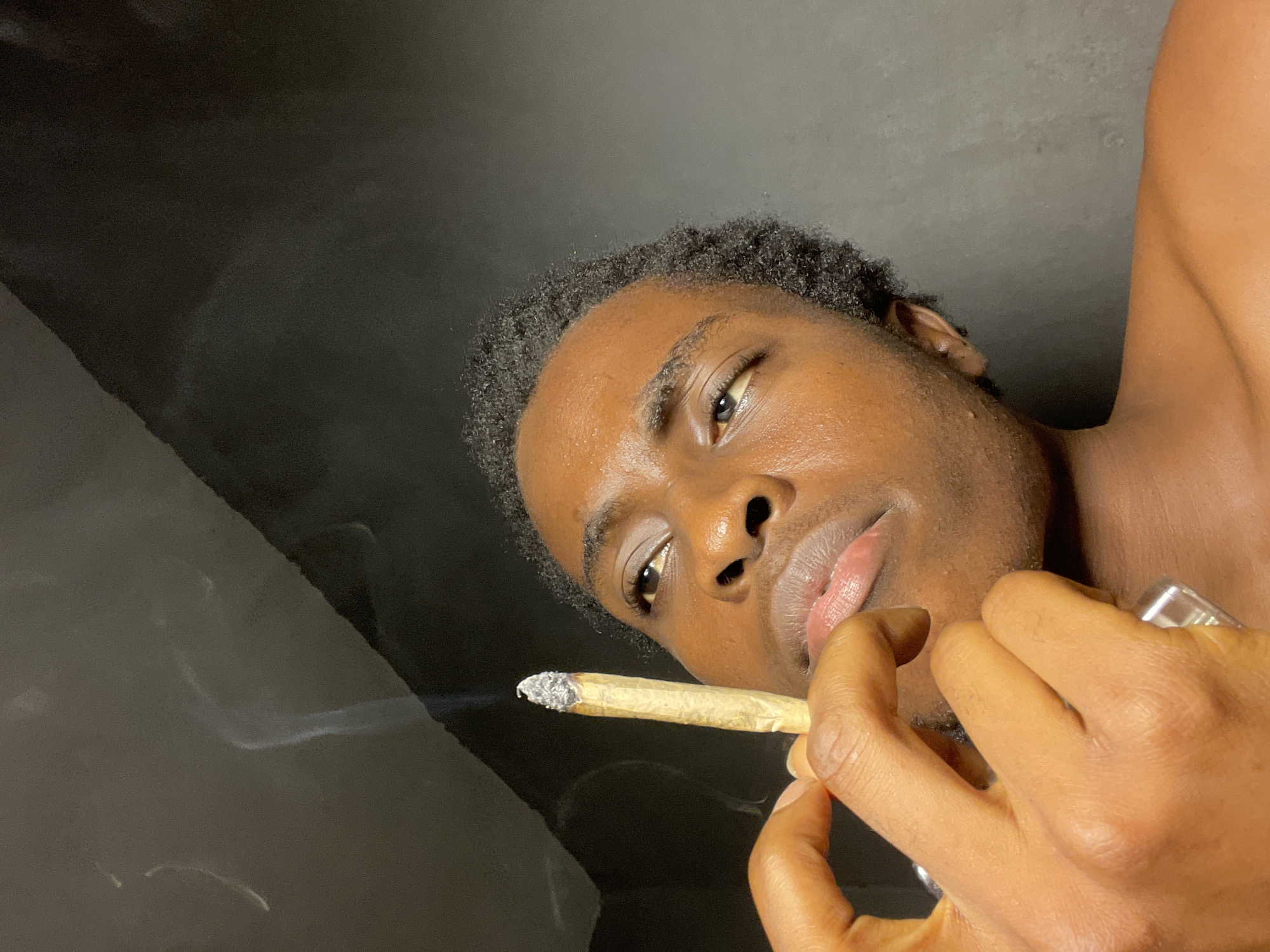 A medium shot depicts a young, dark-skinned male from a low angle, holding a lit cigarette between his fingers and near his lips. Smoke is emanating from the cigarette. He has short, dark hair and appears to be shirtless. The background is dark and indistinct. There are no other visible subjects or objects. There is no other information to suggest this image was taken in Nkpor, Nigeria.