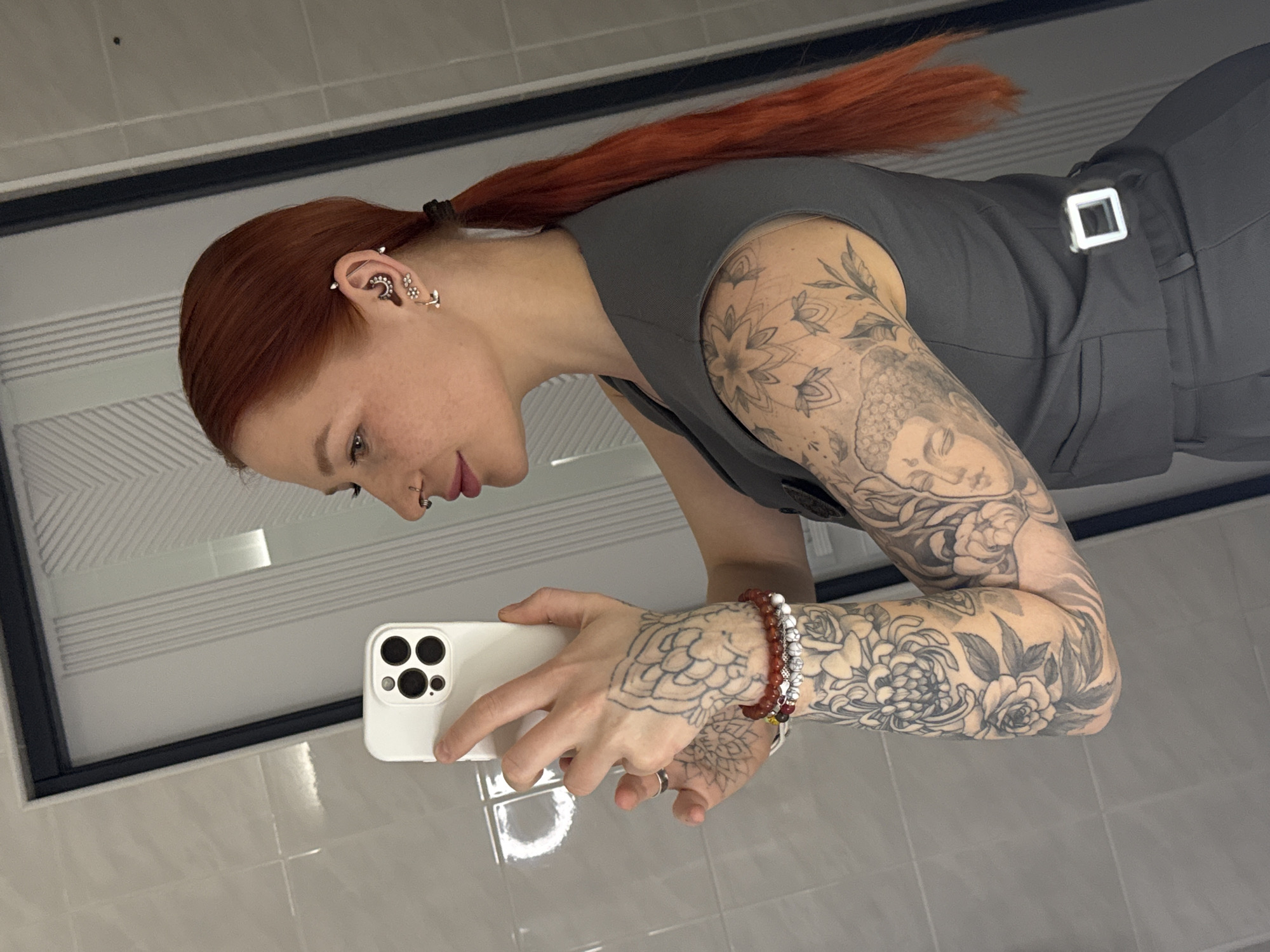 A woman with red hair in a ponytail is reflected in a mirror. She is wearing a gray top and pants with a square buckle. She is holding a white smartphone with a multi-camera system in her hands. The woman has multiple ear piercings and extensive tattoos on her arms, including floral designs and a depiction of a Buddha. She also wears beaded bracelets on her wrist. The background consists of light gray tiled walls. The location is not discernible as Ovoshchnoy, Russia.