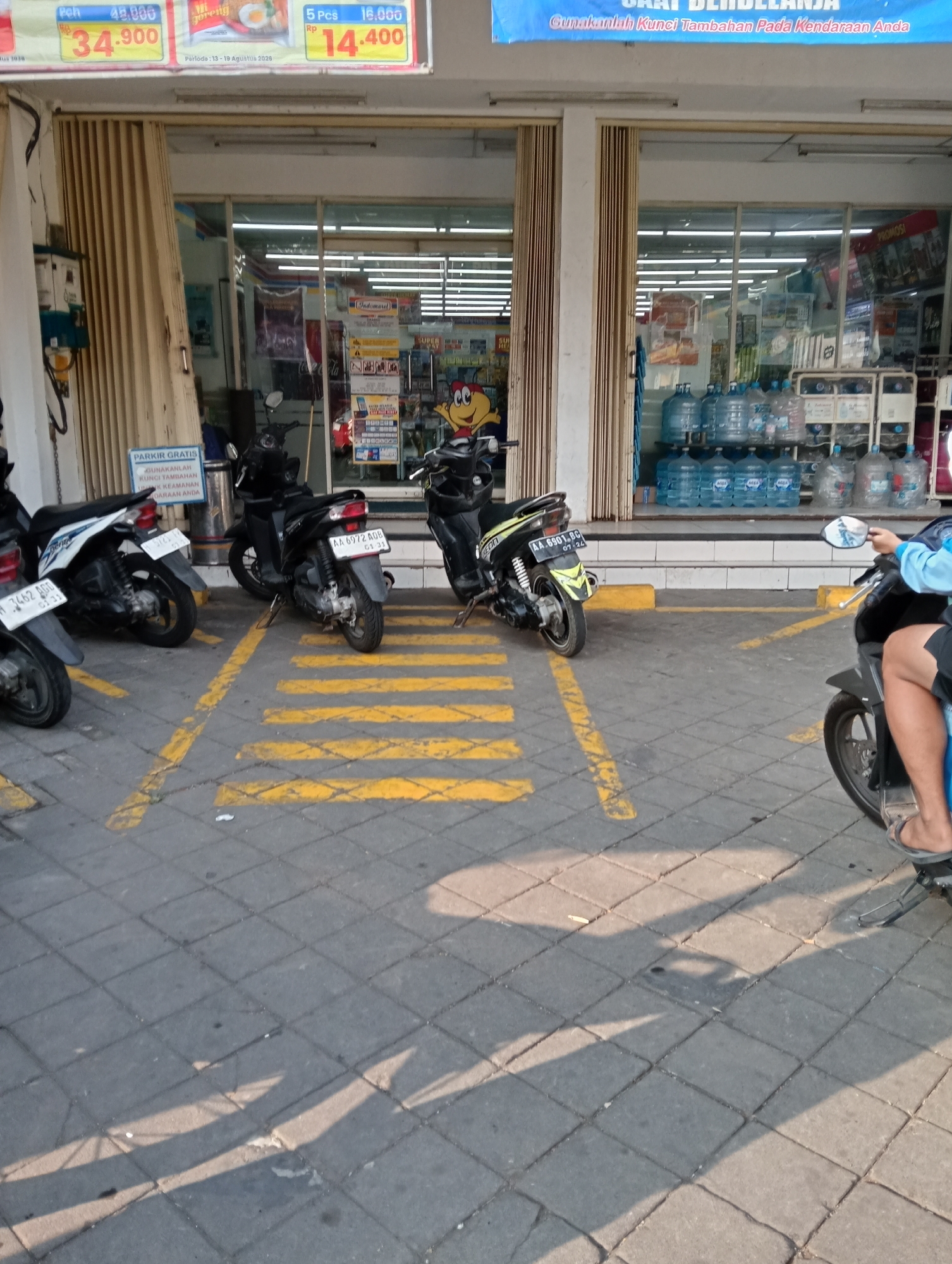 A side view shows several motorcycles parked in designated yellow striped parking spots outside a minimarket in Grabag, Indonesia. The minimarket's entrance features a folding metal door and large glass windows displaying shelves stocked with bottled water and other products. A sign above the entrance advertises "SAAT BERBELANJA" (When Shopping). A person is seated on a motorcycle in the foreground, facing away from the camera. The pavement is made of square gray tiles.