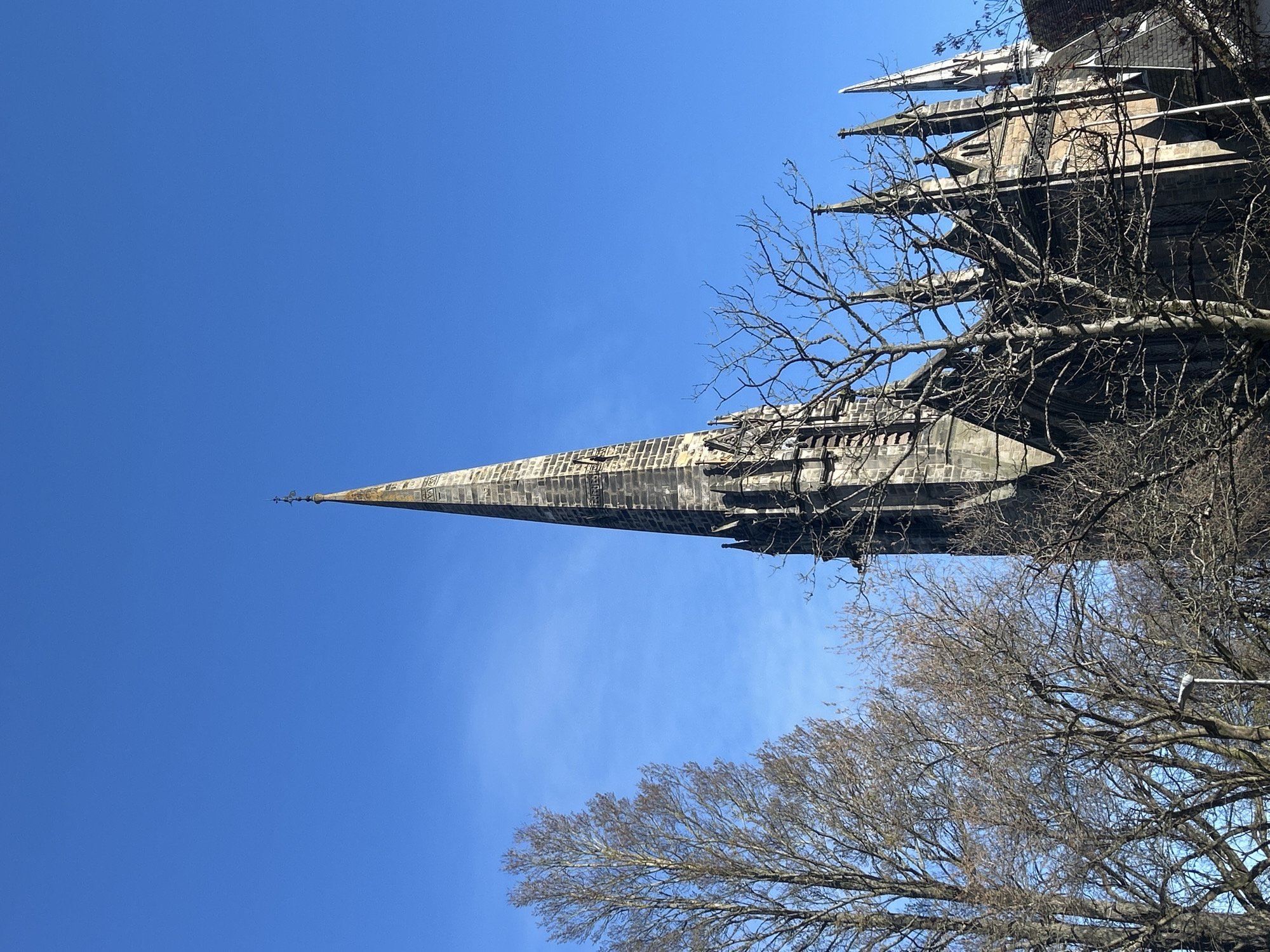 The image, taken in Dundee, United Kingdom, presents a low-angle view of a prominent, tall stone church spire and the upper portions of a Gothic-style building set against a clear, brilliant blue sky. The slender spire, adorned with intricate stonework, tapers dramatically towards a small finial or cross at its summit, dominating the left side of the frame.
To the right, the detailed upper architecture of the church is visible, featuring multiple sharp pinnacles, buttresses, and ornate carvings typical of Gothic design. Interspersed throughout the scene, and particularly in the foreground, are numerous bare, deciduous tree branches, completely devoid of leaves. Their intricate, dark silhouettes contrast sharply with the bright sky and the pale stone of the building, strongly suggesting a winter or early spring season.
The sky is a uniform, deep blue, indicating a bright and sunny day with no clouds. The sunlight illuminates the stone, revealing its texture and architectural depth. There are no people or specific activities depicted. The overall impression is one of historical grandeur and architectural beauty against a crisp, clear sky. No discernible text is visible on the building or in the surroundings.
