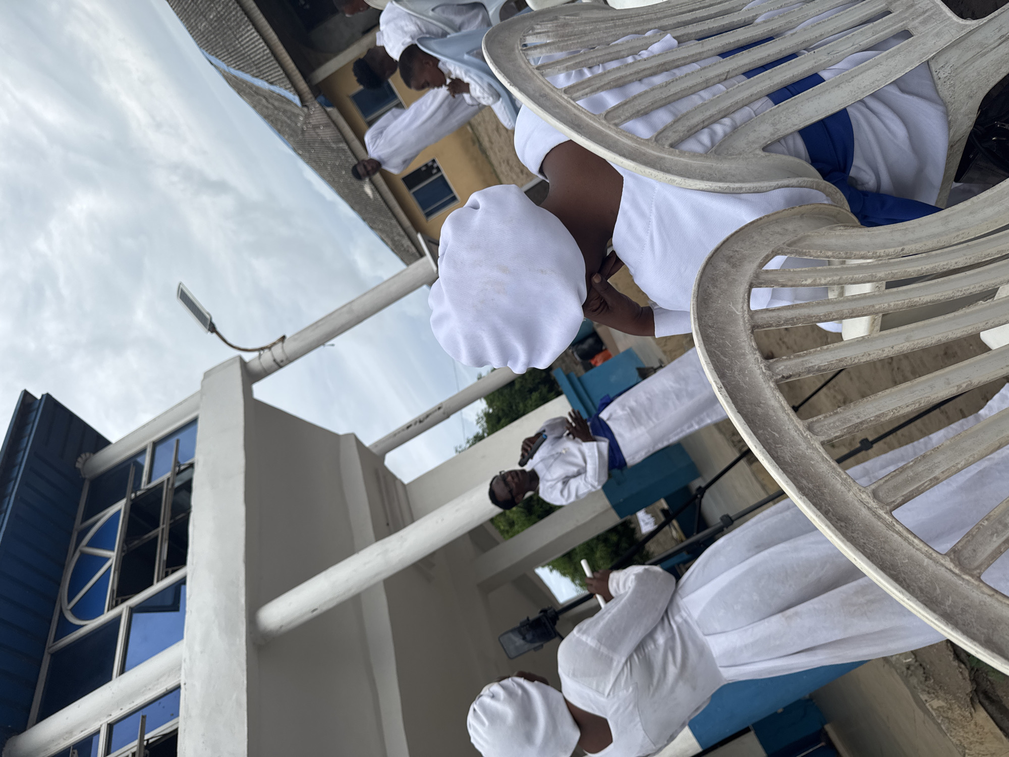The image shows a group of people wearing white robes and caps, possibly attending a religious ceremony or event. They are positioned outside a building with blue-framed windows, and the scene appears to be set in Badagry, Nigeria. People are seated in white plastic chairs. The overall environment seems to be outdoors under cloudy sky.