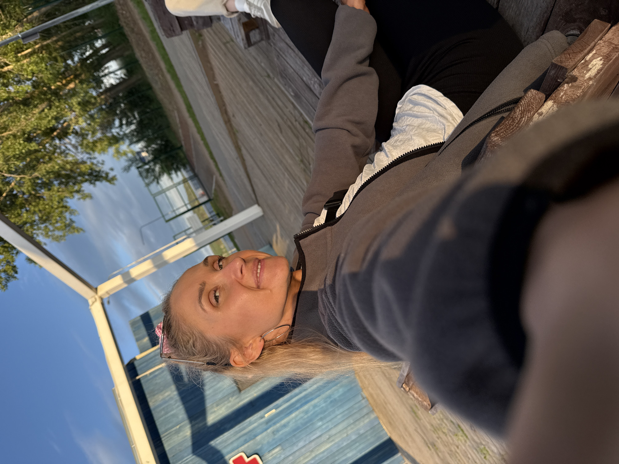 An outdoor, eye-level selfie captures a fair-skinned woman with blonde hair and sunglasses perched on her head, smiling towards the camera. She wears silver hoop earrings, a grey fleece jacket over a white top, and black leggings, sitting on a weathered wooden bench or deck. The setting appears to be a waterfront promenade or park in the city of Chaikovsky, Russia, during golden hour, evidenced by warm, directional light.
To the left, a large blue building with horizontal siding features a partial red cross symbol. Overhead, white structural beams of a canopy or shelter are visible against a blue sky with scattered clouds. The ground is composed of wooden planks. To the right and in the background, a line of tall green trees borders a paved or gravel path. Beyond the trees, a sports court or paved area is visible, followed by a body of water, consistent with the Kama River or a reservoir near Chaikovsky. A metal fence and a utility pole are also present in the distant background. The woman's right hand is partially visible in the lower foreground, suggesting she is holding the camera for the selfie.