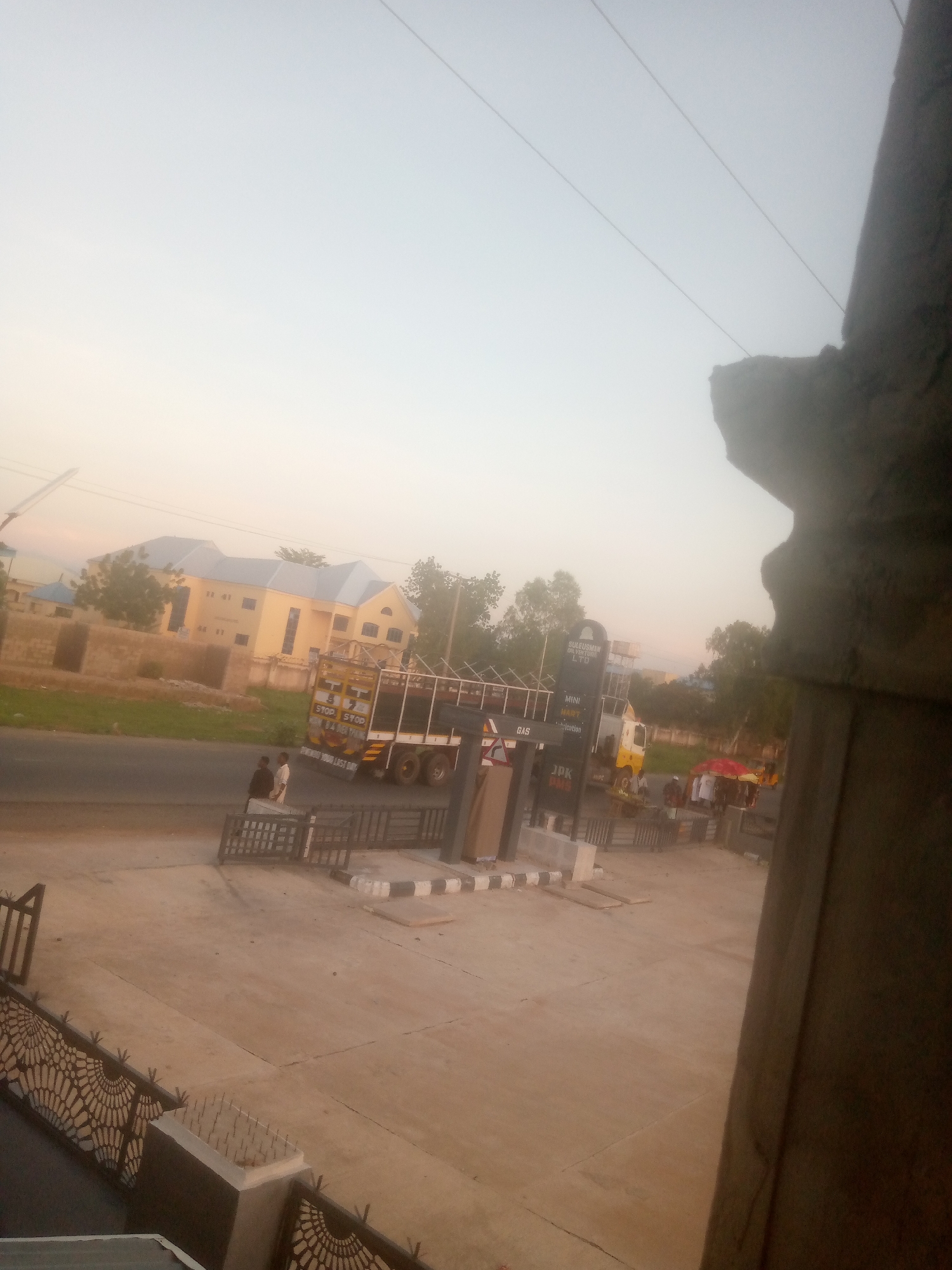 This image captures an elevated perspective of a roadside scene in Jalingo, Nigeria, under hazy daylight conditions, possibly early morning or late afternoon. The sky displays a pale blue-white gradient with a yellowish hue near the horizon, crisscrossed by several utility wires.
In the immediate foreground on the left, a dark, ornate metal fence with a radial pattern is visible, adjacent to a concrete pillar topped with anti-climb spikes. The ground in this section is a light-colored, expansive paved surface, likely concrete. A blurred, dark structural element occupies the right side of the foreground, suggesting it is part of the structure from which the photograph was taken.
The midground features a paved road running horizontally across the frame. A prominent yellow and black flatbed truck, equipped with a metal cage structure, is positioned on the road. The truck's rear displays "STOP STOP" and partially visible text that reads "REMEMBER YOUR LAST DAY." Two individuals stand on the pavement directly beside the truck. Further down the road, a gas station is identifiable by a tall sign listing "SULEIMAN OIL & GAS LTD," "MINI MART," "GAS," and "JDK DMS." Another lighter-colored truck or van is parked near the gas pumps. Several people are visible in the vicinity of the gas station, including one standing beneath a red umbrella. Black and white striped concrete barriers delineate the gas station's forecourt.
The background primarily consists of a large, multi-story building painted yellow with a gray roof, situated behind the road and surrounded by numerous green trees and sparse vegetation. Other smaller buildings are faintly visible in the distance.