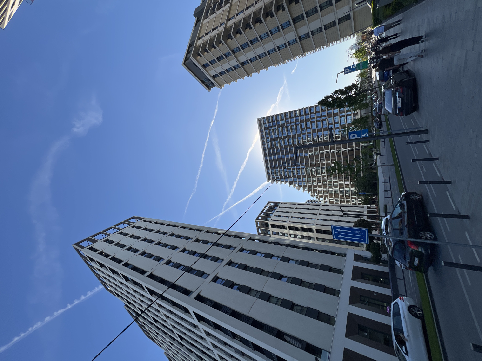 The image, captured from a low angle with a rotated perspective, showcases a modern urban environment on a bright, sunny day in Belgrade, Serbia. Several contemporary high-rise buildings with distinctive geometric facades and balconies dominate the frame, stretching towards a clear blue sky. The bright sun is partially obscured by one of the buildings, creating a radiant glow and lens flare, while multiple white contrails streak across the sky.
At street level, a paved road with white markings runs parallel to the buildings. A line of parked cars is visible, including a dark sedan with a Serbian license plate displaying "AP 0244 JB" and a white hatchback. Several people are seen walking on the sidewalk alongside the parked vehicles. Street furniture includes blue parking signs marked with a white 'P' symbol, and another blue sign depicting upward and downward arrows, along with a bicycle symbol, indicating a two-way street or a specific lane designation. A dark utility line or cable crosses the upper left portion of the frame. The overall impression is one of a bustling city scene under clear skies.