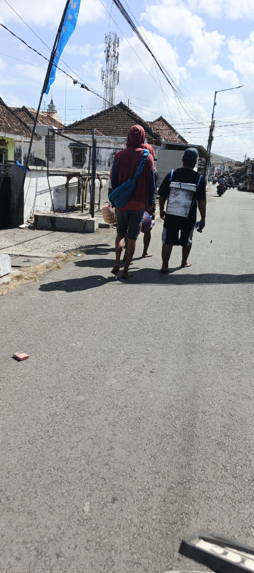 Two men and a child are walking on a paved road in Puger, Indonesia, under a bright, cloudy sky. The man on the left wears a maroon hooded sweatshirt and gray shorts, carrying a blue bag over his shoulder and a woven basket in his hand. To his right, a child in a red shirt and patterned shorts walks behind him. The man on the right wears a black t-shirt and patterned shorts, with a white rectangular object strapped to his back like a backpack. Both men are barefoot. In the background, a cell tower rises above a row of traditional tiled-roof houses, with power lines crisscrossing overhead. The road shows tire marks and scattered debris, including a red rectangular object near the foreground. To the right, a street lamp is visible, and a few motorcycles are parked in the distance, suggesting a market or residential area. The shadows of the individuals are cast long on the asphalt, indicating it is likely midday.