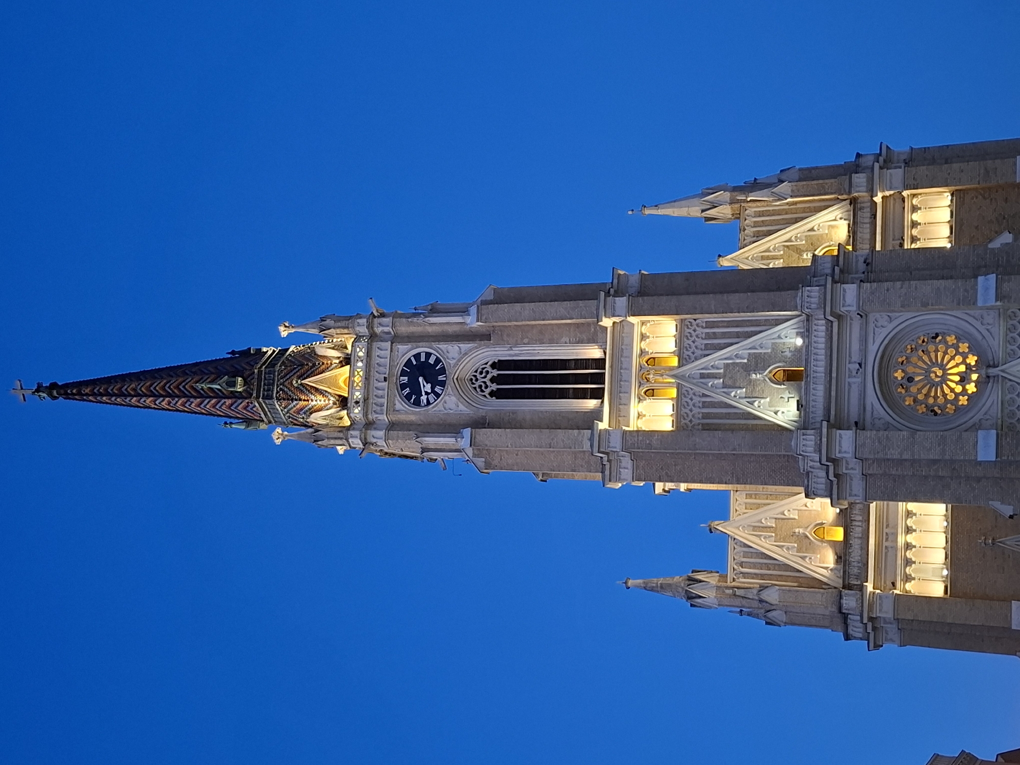 The image depicts the Church of the Name of Mary in Novi Sad, Serbia, taken at dusk or night. The Neo-Gothic church's illuminated steeple is the focal point against a dark blue sky. The steeple features a clock, ornate stonework, and a cross at its peak. The architectural details, including rose windows and lancet arches, are visible. There are no people visible in the image.
