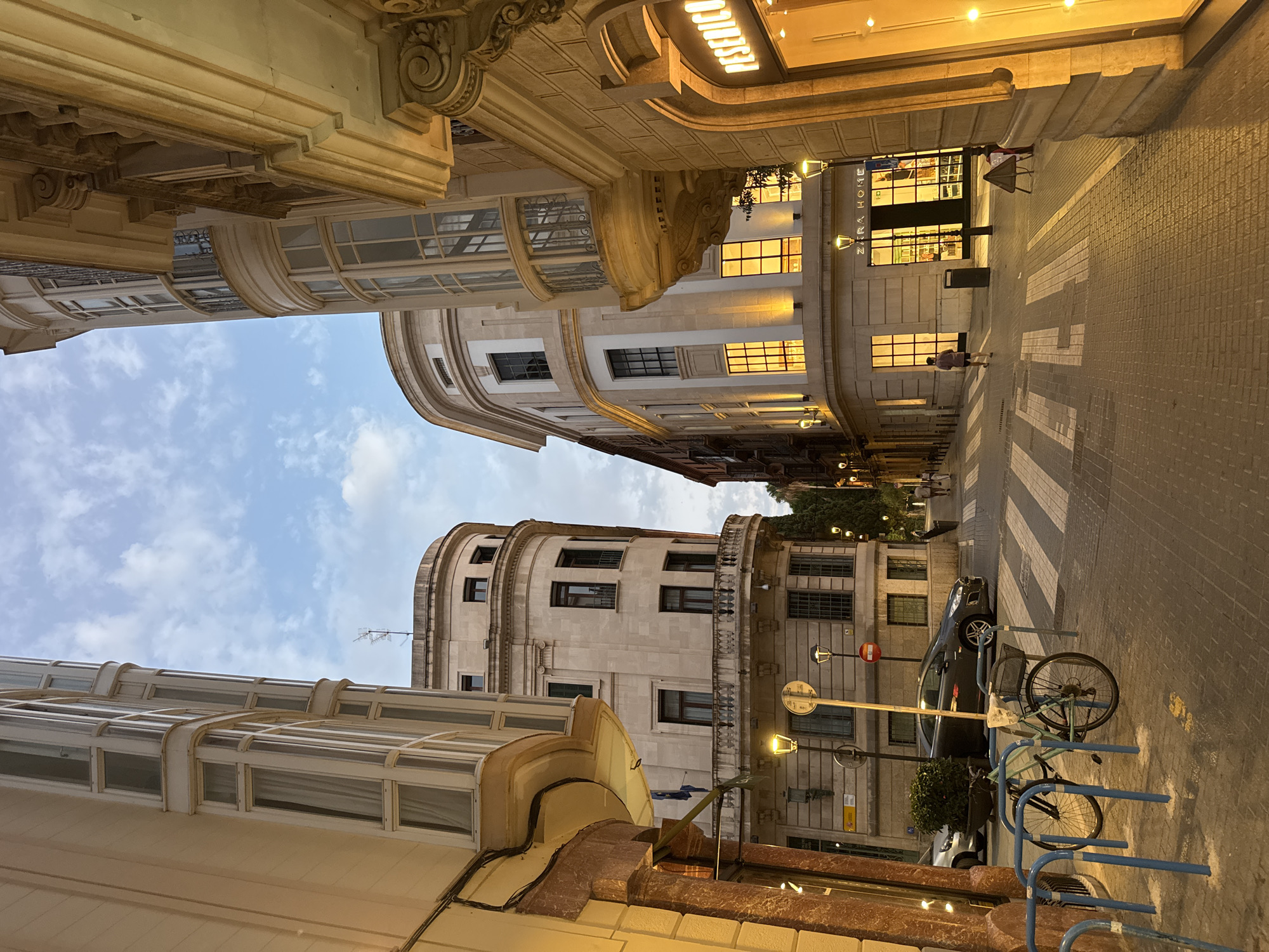 The image depicts a street scene in Palma, Spain, likely during the evening or early morning given the artificial lighting and twilight sky. The perspective is from a low angle, looking down a cobblestone street lined with multi-story buildings.
On the left, a light-colored building features a prominent bay window structure with multiple panes. Its facade is a warm, sandy color, and the ground floor has a narrow storefront. In the foreground, a blue bicycle rack holds two bicycles. One is a light blue cruiser with a basket and what appears to be a newspaper inside. The other bicycle is partially obscured.
Across the street, a pedestrian crossing with white stripes is visible. Several cars are parked along the side of the road, and a dark-colored car is positioned near the crossing. Two streetlights, one with a circular sign, illuminate the scene.
Dominating the center of the image is a large, beige building with rounded architectural features and ornate balconies. Its windows on the upper floors are illuminated with a warm, orange light, suggesting interior lighting. The ground floor of this building appears to be a retail establishment labeled "ZARA HOME."
To the right, another building with a decorative facade is illuminated by soft yellow light emanating from its large storefront windows. A sign with the word "PESERICO" is visible above this storefront.
A few people are present in the scene: one individual is walking away from the camera on the right side of the street, and another figure is further down the street, also walking away. The sky above is a pale blue with scattered clouds. The overall atmosphere is calm and somewhat serene, with the urban architecture and street elements defining the environment.