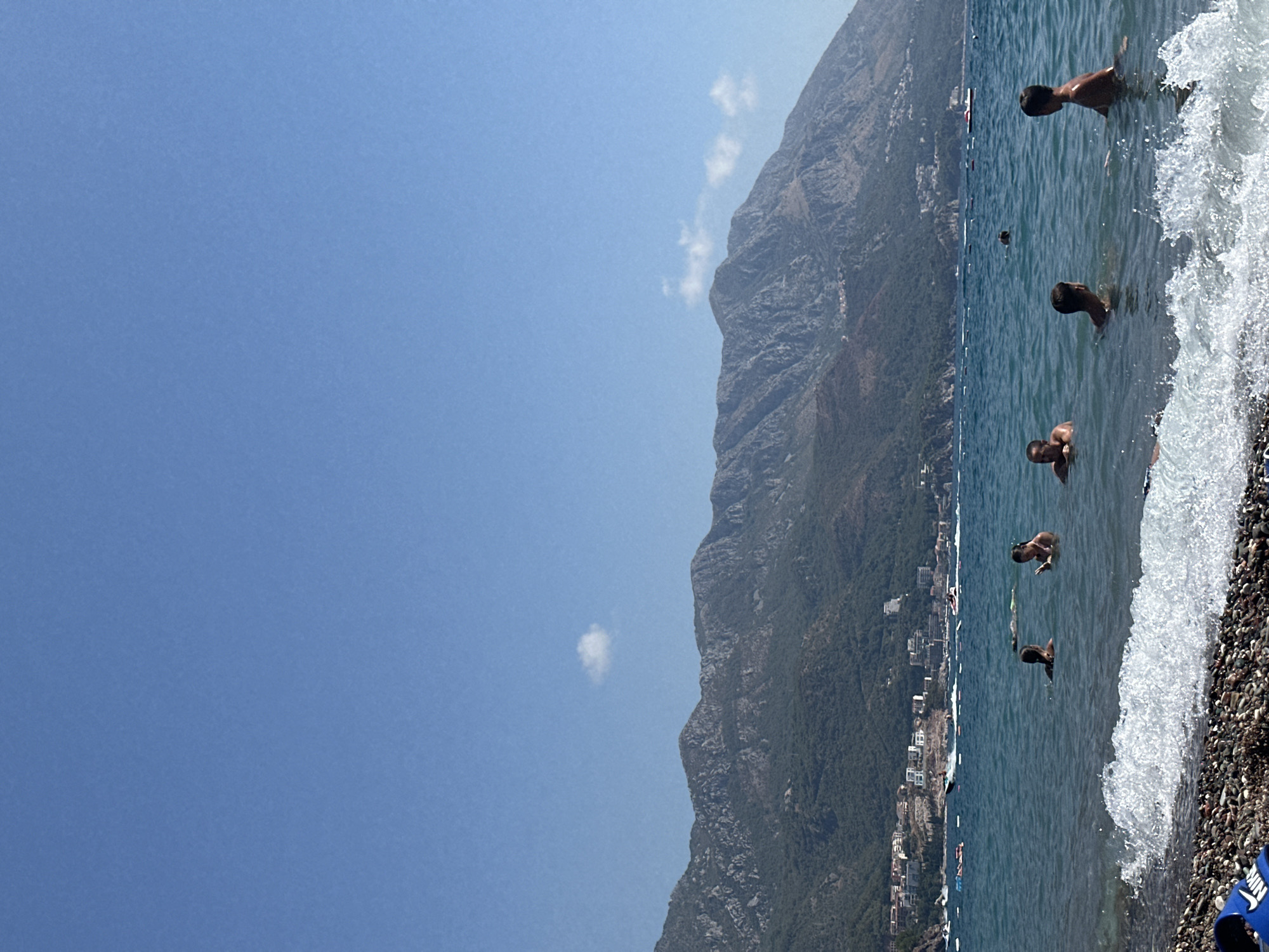 The image depicts a sunny day at a beach in Boreti, Montenegro. In the foreground, the rocky shore is visible with waves gently lapping at the pebbles. The water is a deep blue, and several people are swimming and wading. Further out, boats of various sizes are scattered across the sea, and in the background, a lush, green mountainside rises, dotted with buildings that form part of the town of Boreti. The sky is a clear, bright blue with a few wispy clouds.