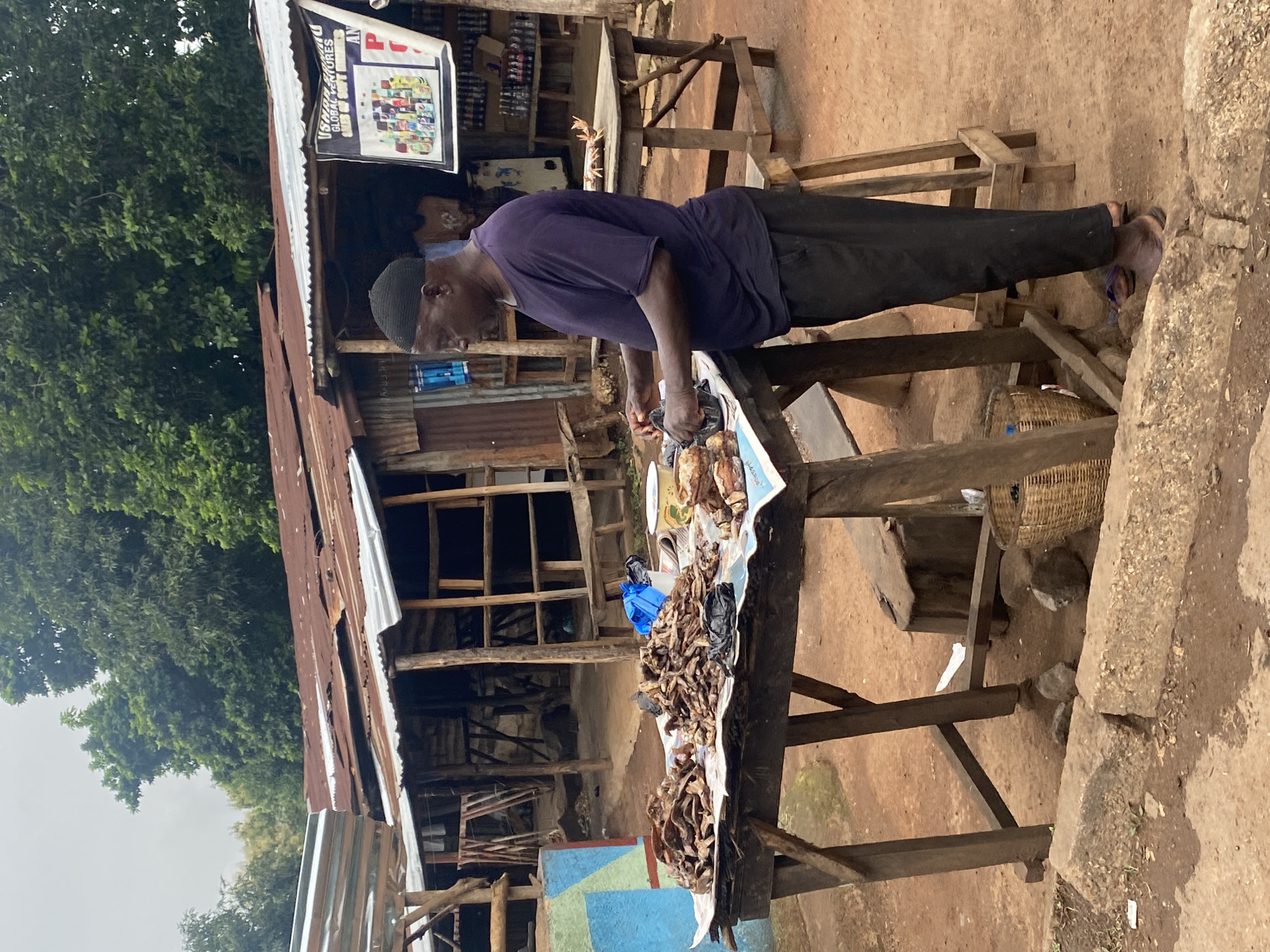 A man is standing at a wooden table in an outdoor market stall in Gidan Waya, Nigeria. He is wearing a purple t-shirt and dark pants, and a dark cap on his head. The man is leaning over the table, which is covered in a white paper displaying various dried food items, possibly fish or meat. He is holding a black plastic bag in his right hand and appears to be sorting or arranging the goods.
The stall itself is a rustic structure made of wood and corrugated metal roofing. Behind the man, shelves are visible, stocked with various items, suggesting this is a general store or a food vendor. A sign hangs above the stall with text and images, possibly advertising products. In the background, lush green trees are visible, indicating a location with vegetation. The ground appears to be dirt, common in many outdoor markets.
The scene suggests an ordinary day at the market, likely during daylight hours given the natural lighting. There are no strong indicators of specific weather conditions, though the overcast sky suggests it might be a cloudy day. The man's expression is neutral, focused on his task. The overall atmosphere is one of everyday commerce in a rural or semi-urban Nigerian setting.